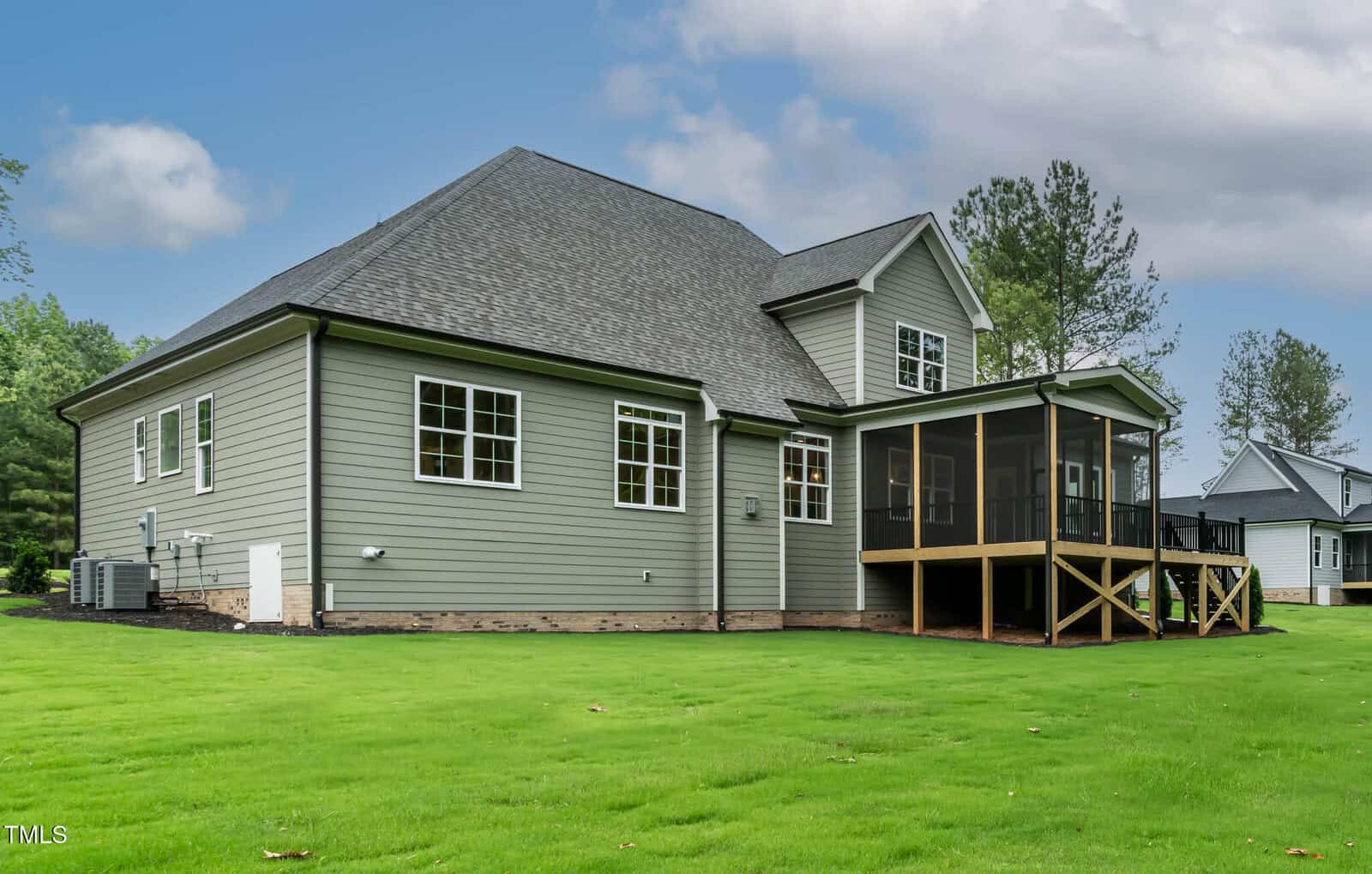 A two-story house with gray siding, white trim, and a screened-in porch. The yard is green and well-maintained, and mature trees and neighboring houses are visible in the background under a partly cloudy sky.