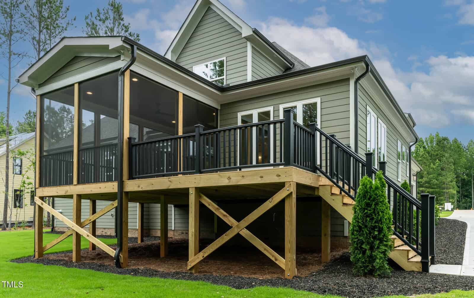A gray house with white trim features a raised wooden deck with black railings and a screened-in porch area. The yard is landscaped with mulch and green grass, and trees are visible in the background.