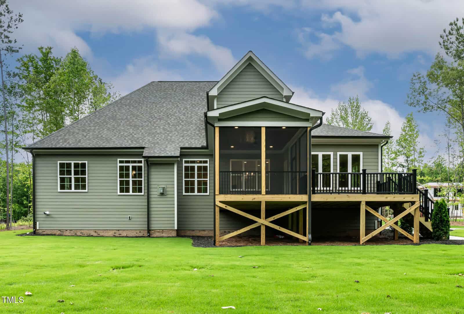 A modern single-story house with gray siding and a dark roof, featuring a screened porch and a raised wooden deck overlooking a large green lawn, surrounded by trees under a partly cloudy sky.