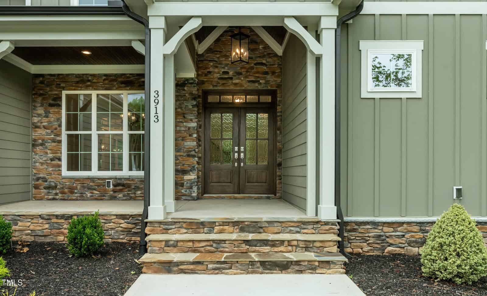 Front entrance of a house with stone and green siding, a covered porch supported by white columns, a double wooden door with glass panels, and steps leading up to the porch. Small shrubs are planted on either side.