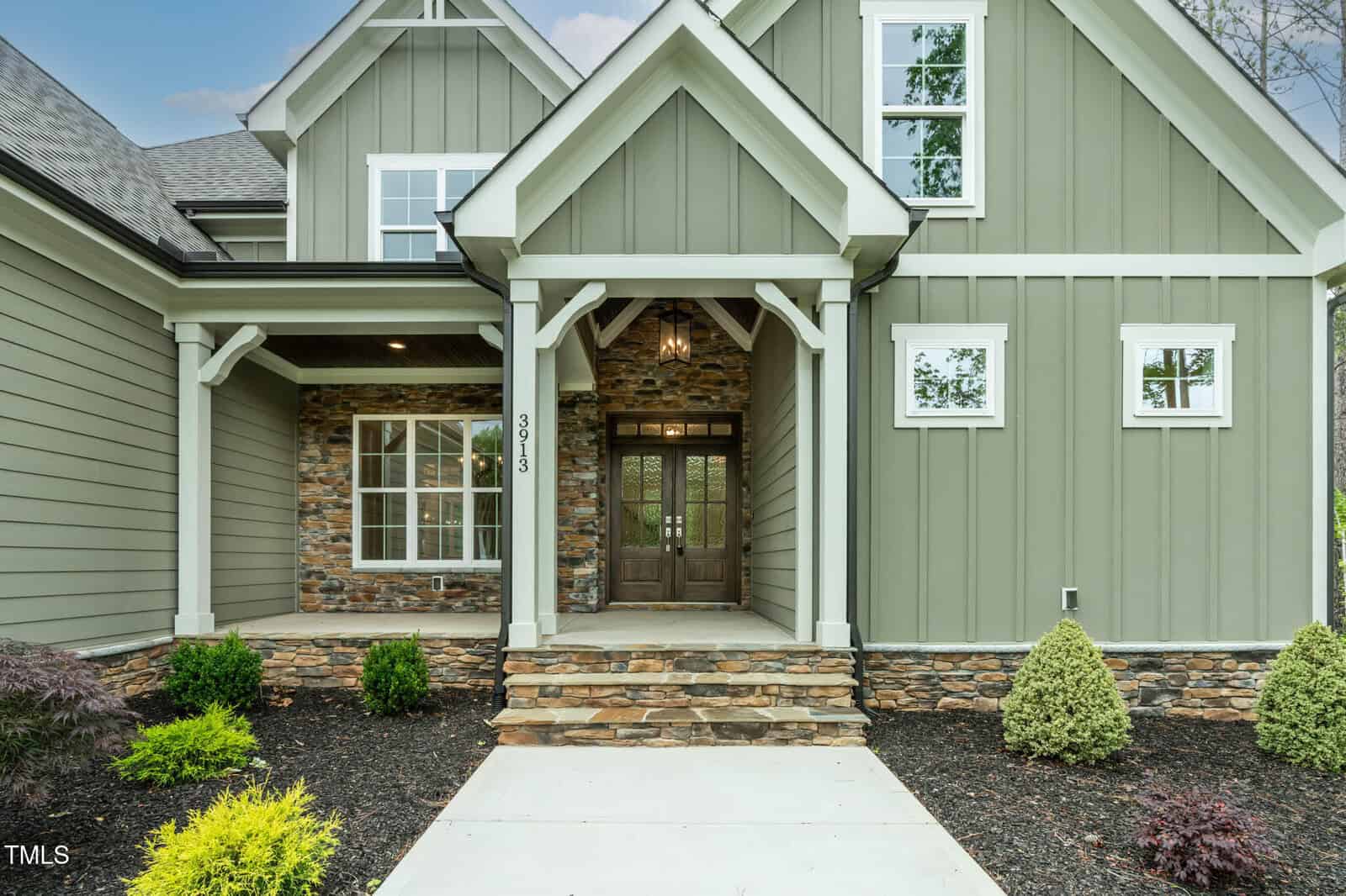 A front view of a modern house with green vertical siding, stone accents, white trim, a covered entryway with wooden double doors, and landscaped shrubs along the walkway.
