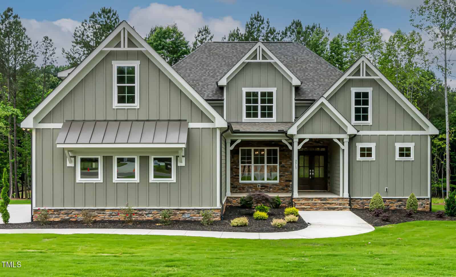 A modern two-story house with gray board-and-batten siding, white trim, stone accents, and large windows, set on a manicured lawn with young landscaping and surrounded by trees.