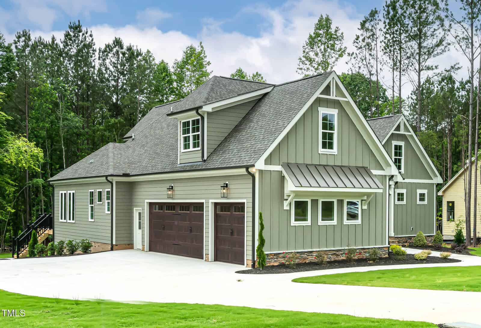 A modern gray house with white trim, two brown garage doors, and a metal awning over a side window. The house is surrounded by green grass, trees, and a concrete driveway.
