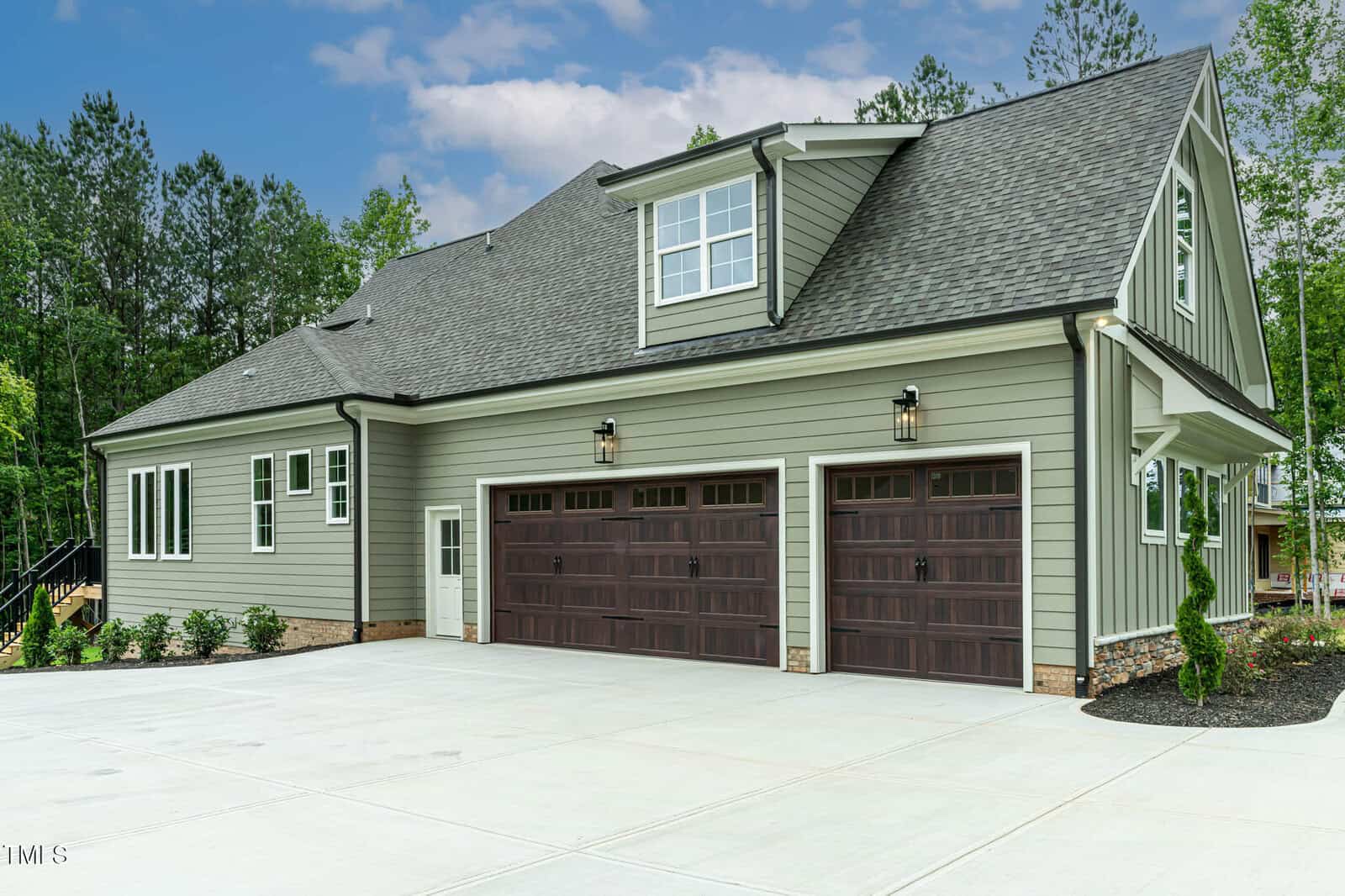 A modern two-story house with green siding and a large driveway. The home features three wooden garage doors, multiple windows, and is surrounded by trees and landscaped greenery.