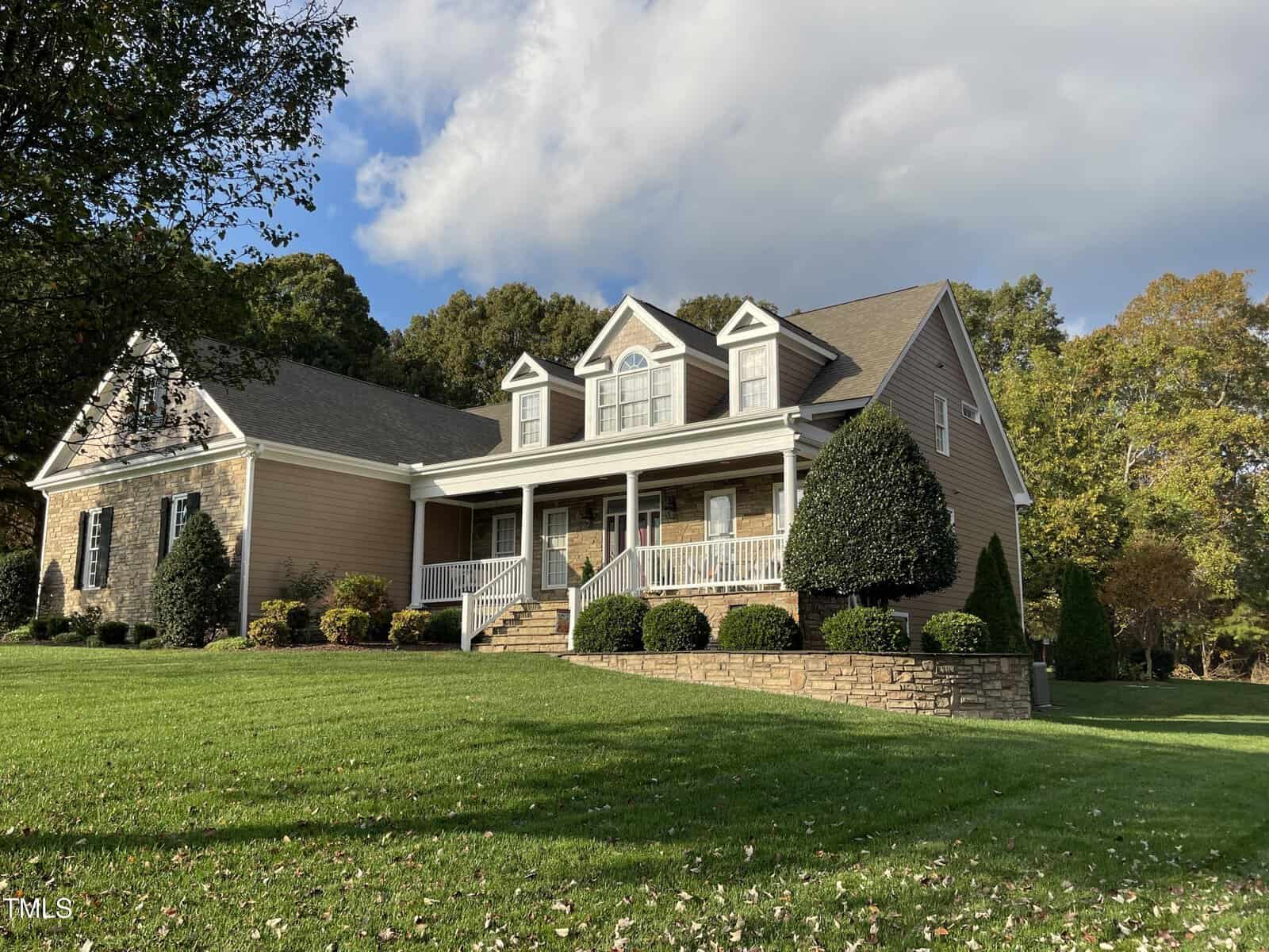 A two-story beige house with white trim, a covered front porch, dormer windows, and manicured shrubs sits on a large lawn with a stone retaining wall and trees in the background on a sunny day.