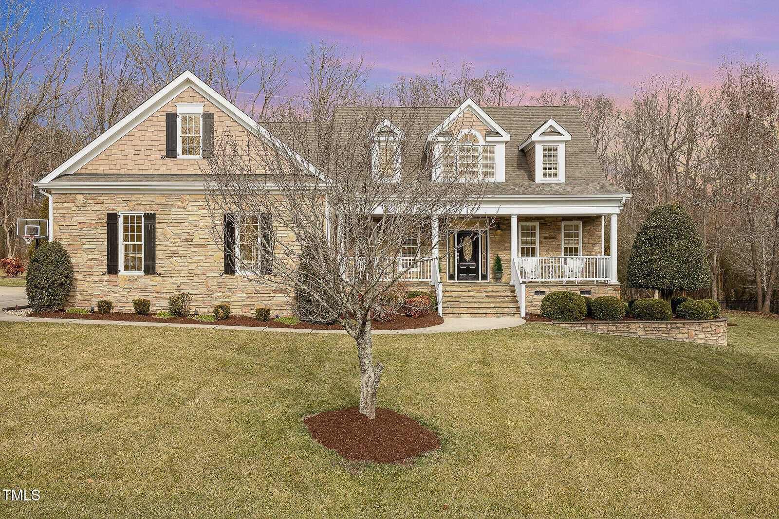 A two-story house with stone and siding exterior, front porch with white railings, trimmed bushes, a leafless tree in the yard, and a basketball hoop on the driveway. Trees without leaves are in the background.