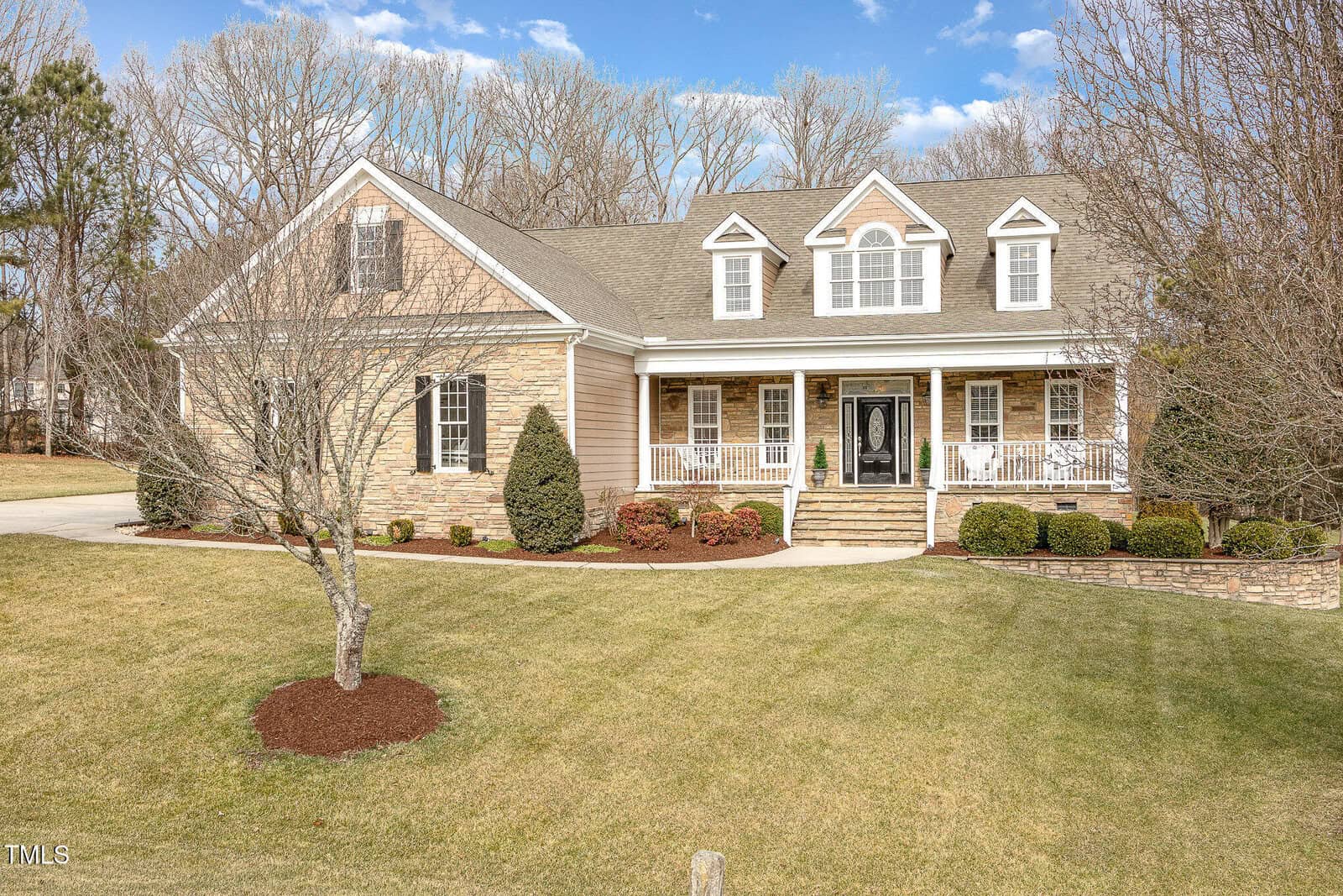 A two-story beige house with stone accents, a covered front porch, dormer windows, and a manicured lawn with trimmed shrubs and leafless trees, set against a backdrop of tall, bare trees and a blue sky.