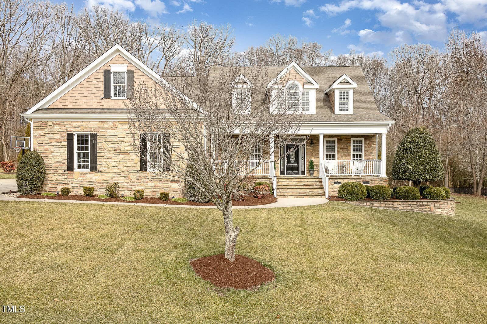 A two-story beige house with stone and siding exterior, white trim, and a covered front porch. The yard is neatly landscaped with trimmed bushes, a tree in front, and lawn, with a wooded area in the background.