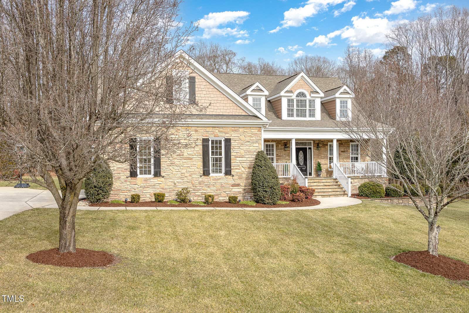 A two-story house with stone and siding exterior, white trim, dormer windows, a covered front porch, and landscaped yard with trees and shrubs on a clear day.