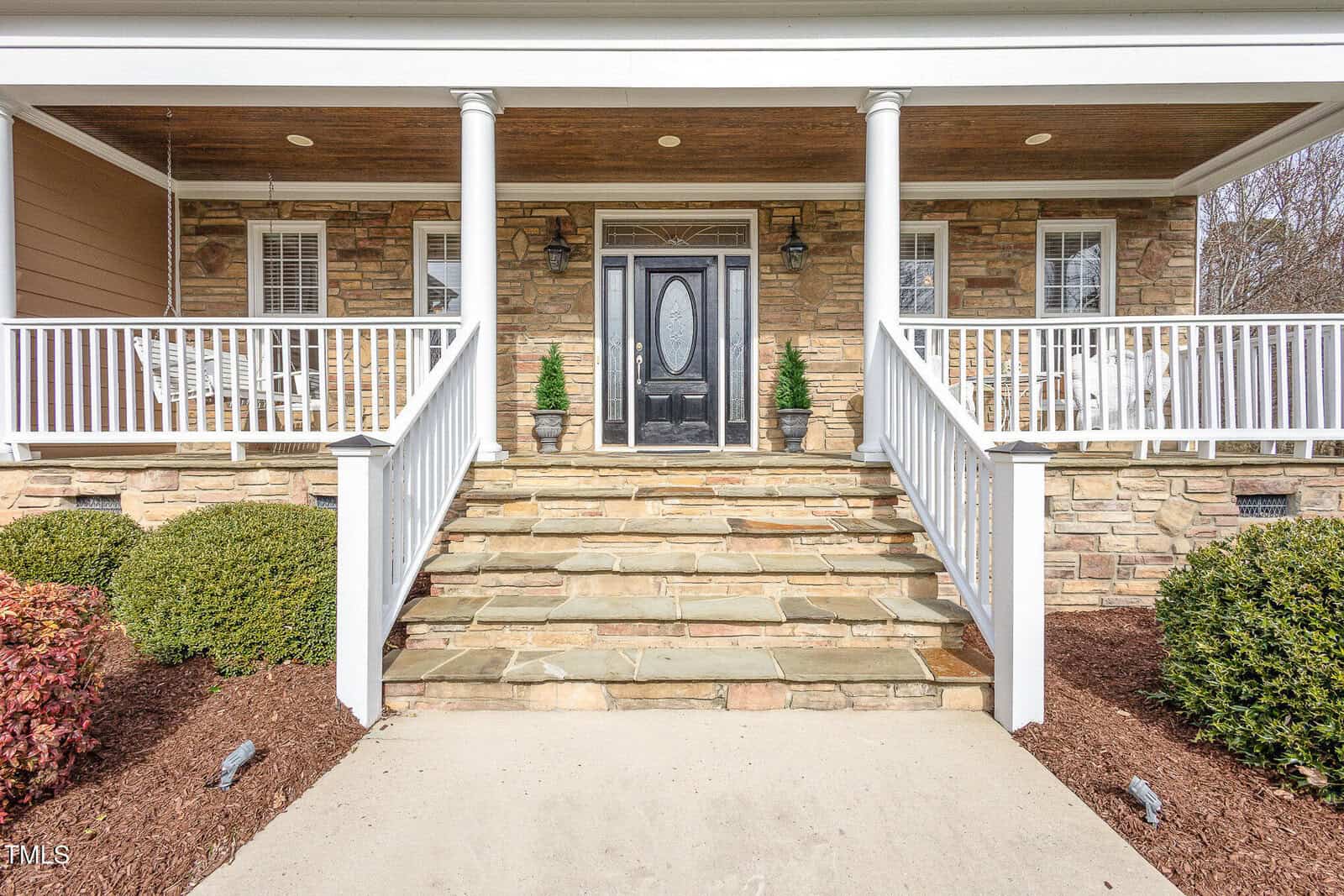 Stone steps lead to a porch with white railings, two white porch swings, a black front door with glass panel, and two potted plants beside the entrance. Shrubs and mulch line the walkway.