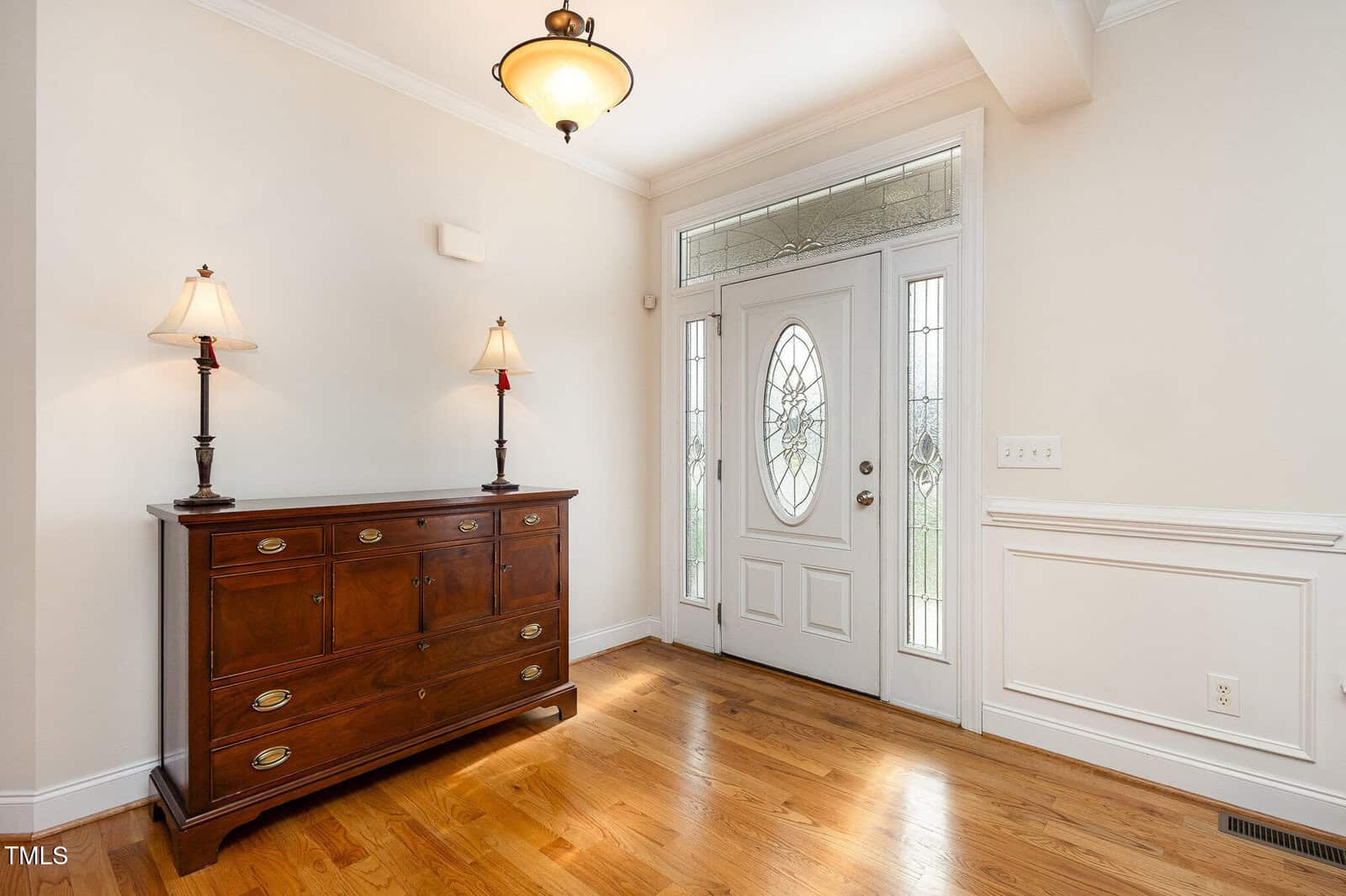 Bright entryway with light wood floors, a white front door with decorative glass panels, a wooden chest with brass handles, and two table lamps. Crown molding and wainscoting add classic detail to the room.