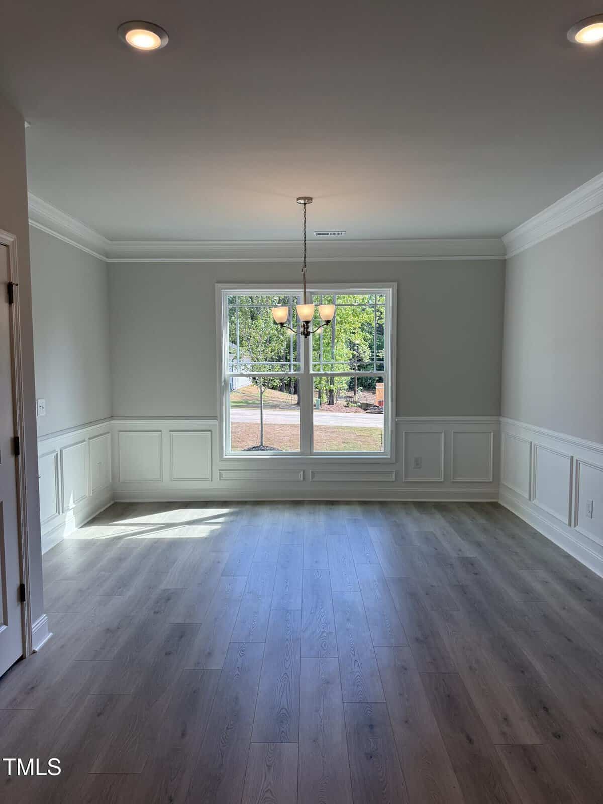 A dining room with light gray walls, white wainscoting, wood-style flooring, a central window, and a five-light chandelier hanging from the ceiling. Sunlight comes in through the large window.