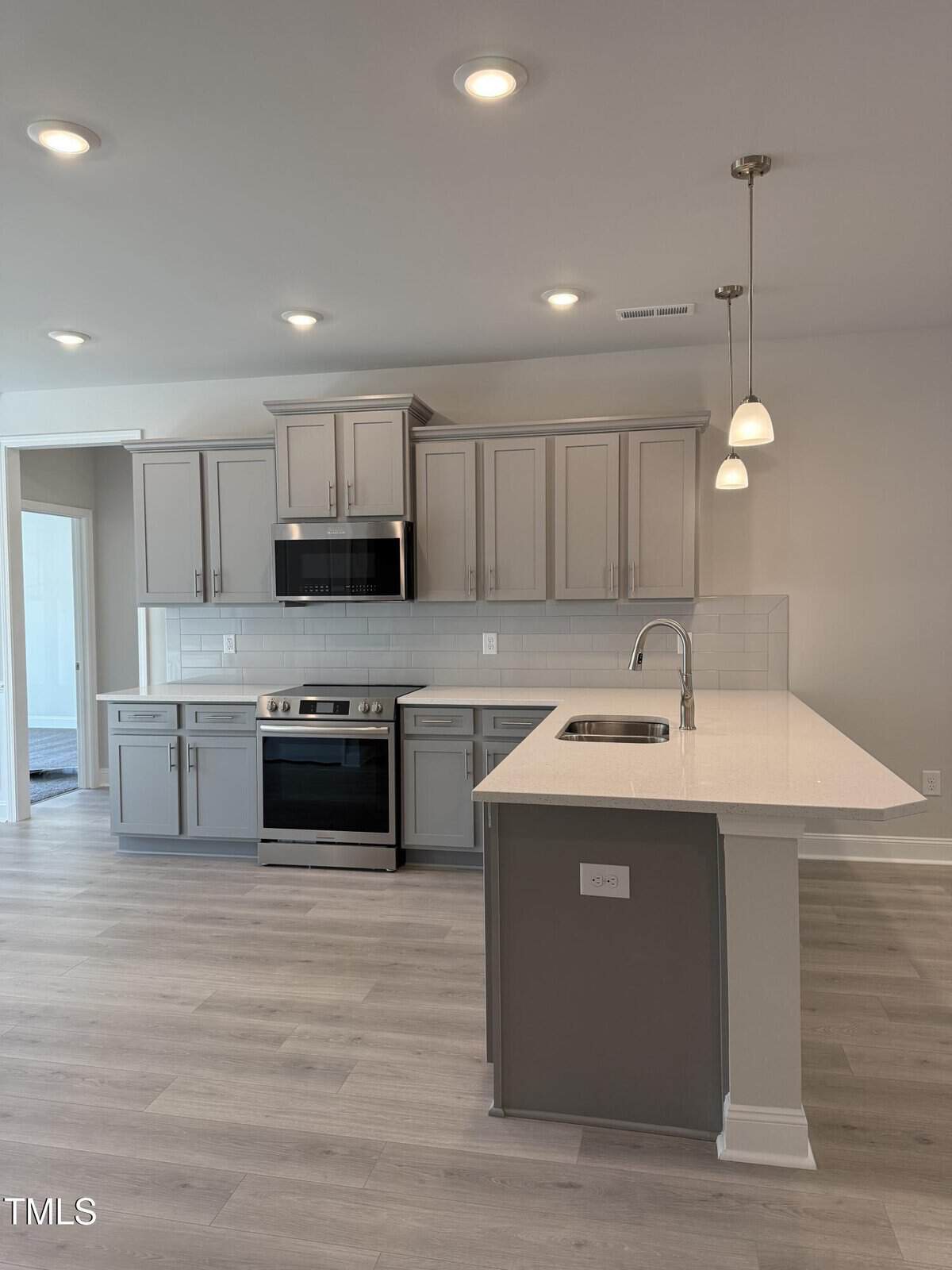 Modern kitchen with gray cabinets, stainless steel appliances, a white tile backsplash, island with sink, white countertops, pendant lights, and light wood flooring.