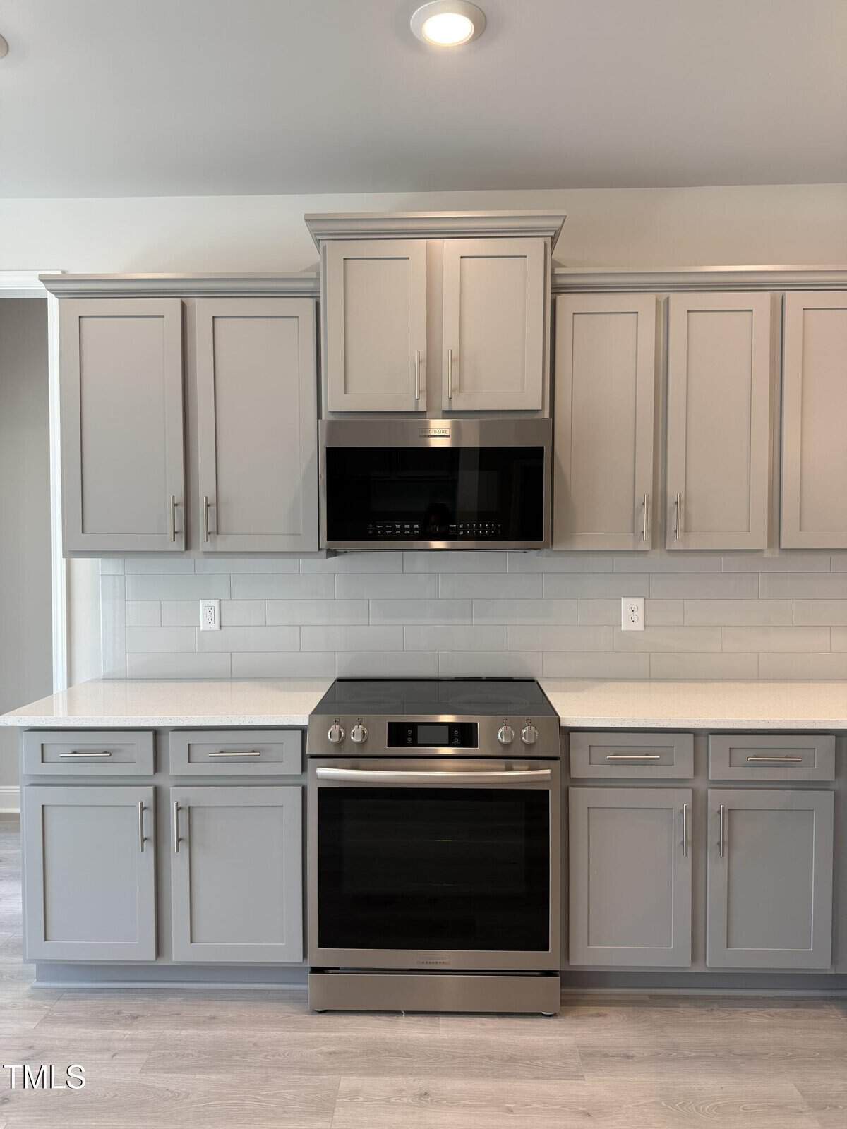 Modern kitchen with light gray cabinets, stainless steel oven, black microwave, white subway tile backsplash, and light countertops. Recessed lighting above and light wood flooring below. No items on the counters.