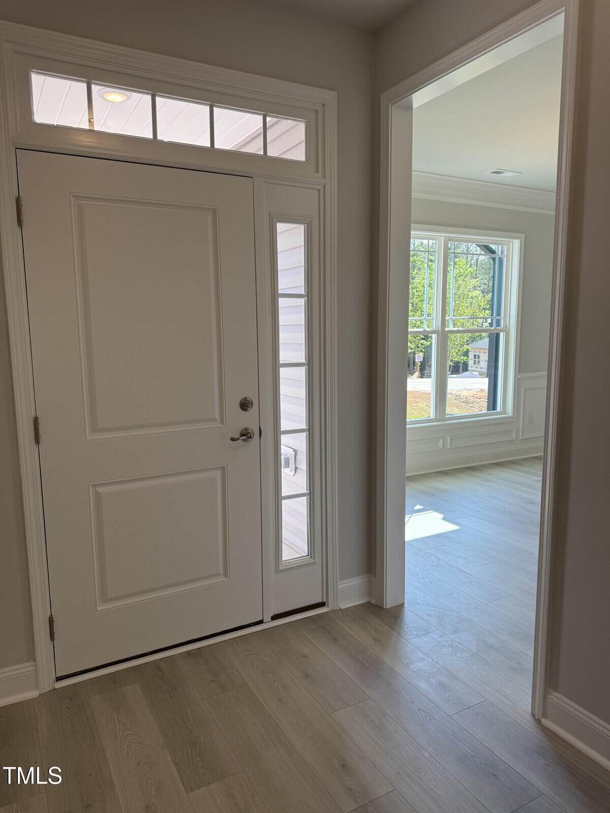 A white front door with a sidelight and window above opens into a hallway with light wood flooring. To the right, there is an open doorway leading to a sunlit room with large windows.
