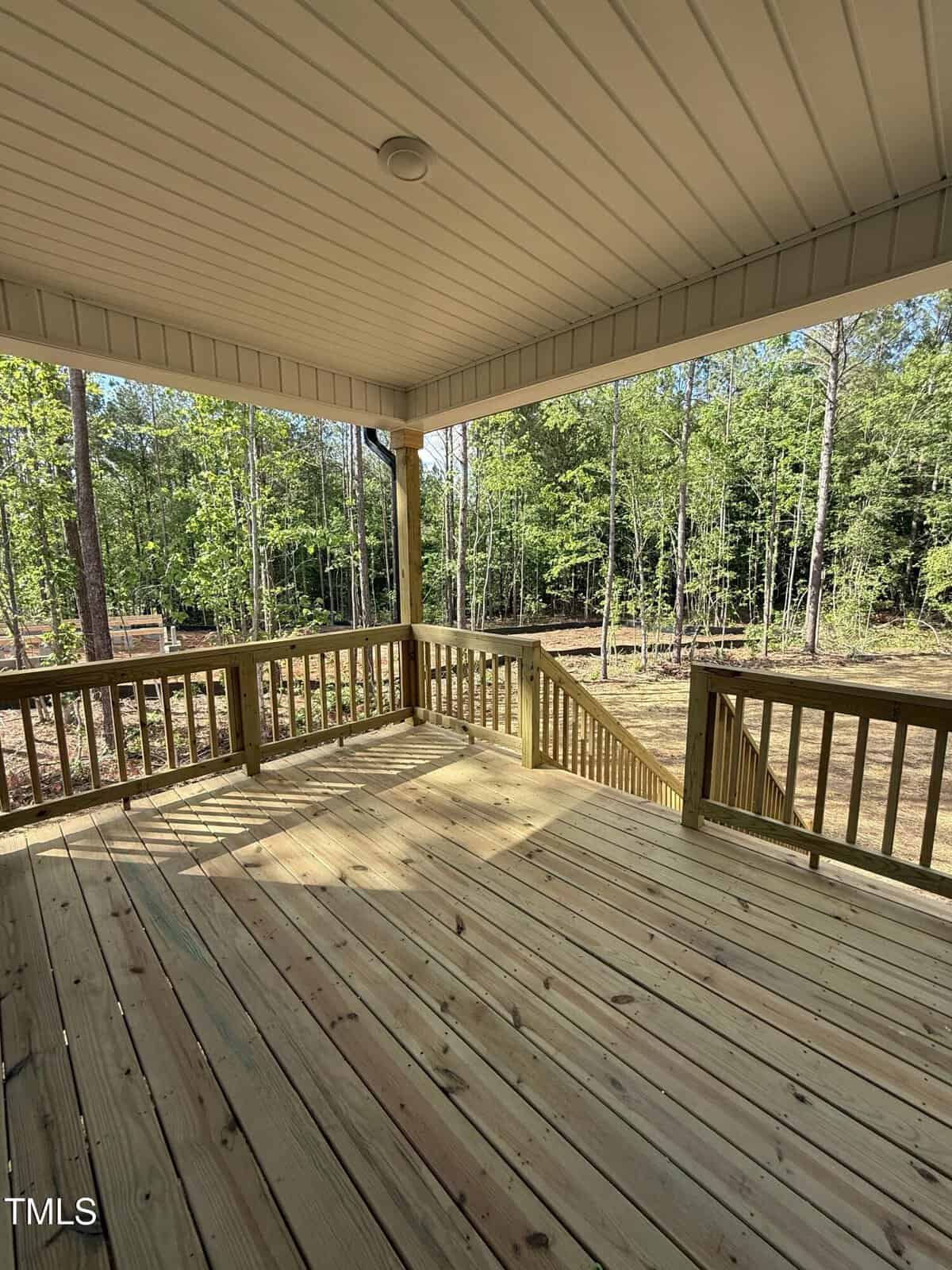 A covered wooden deck with railings and stairs leading down to a grassy area. The deck overlooks a wooded landscape with tall trees. Bright sunlight casts shadows on the deck floor.
