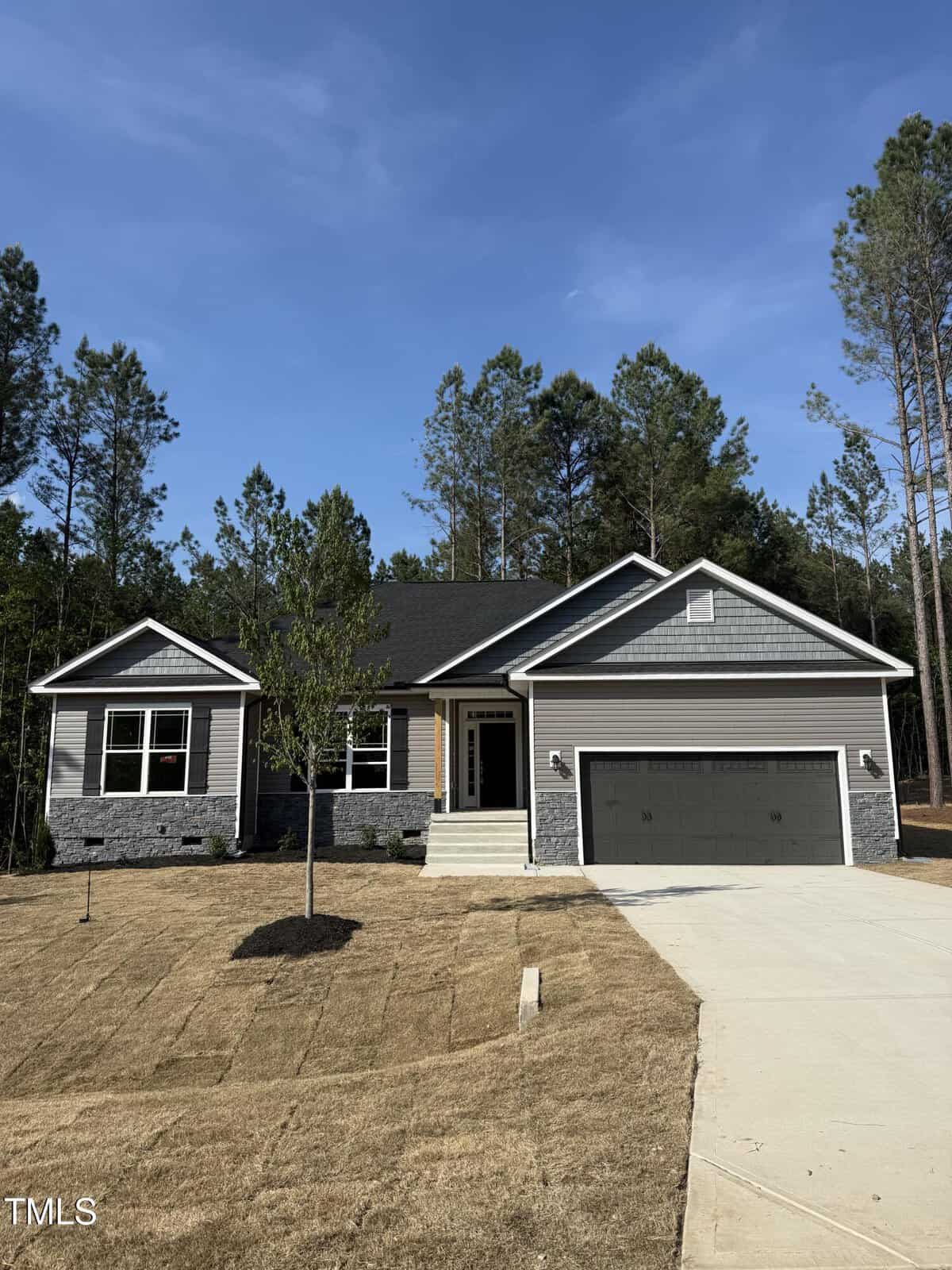 A single-story house with gray siding and stone accents, a dark shingled roof, double garage, and a small front porch. The yard is mostly brown grass with young trees. Tall pine trees line the background.