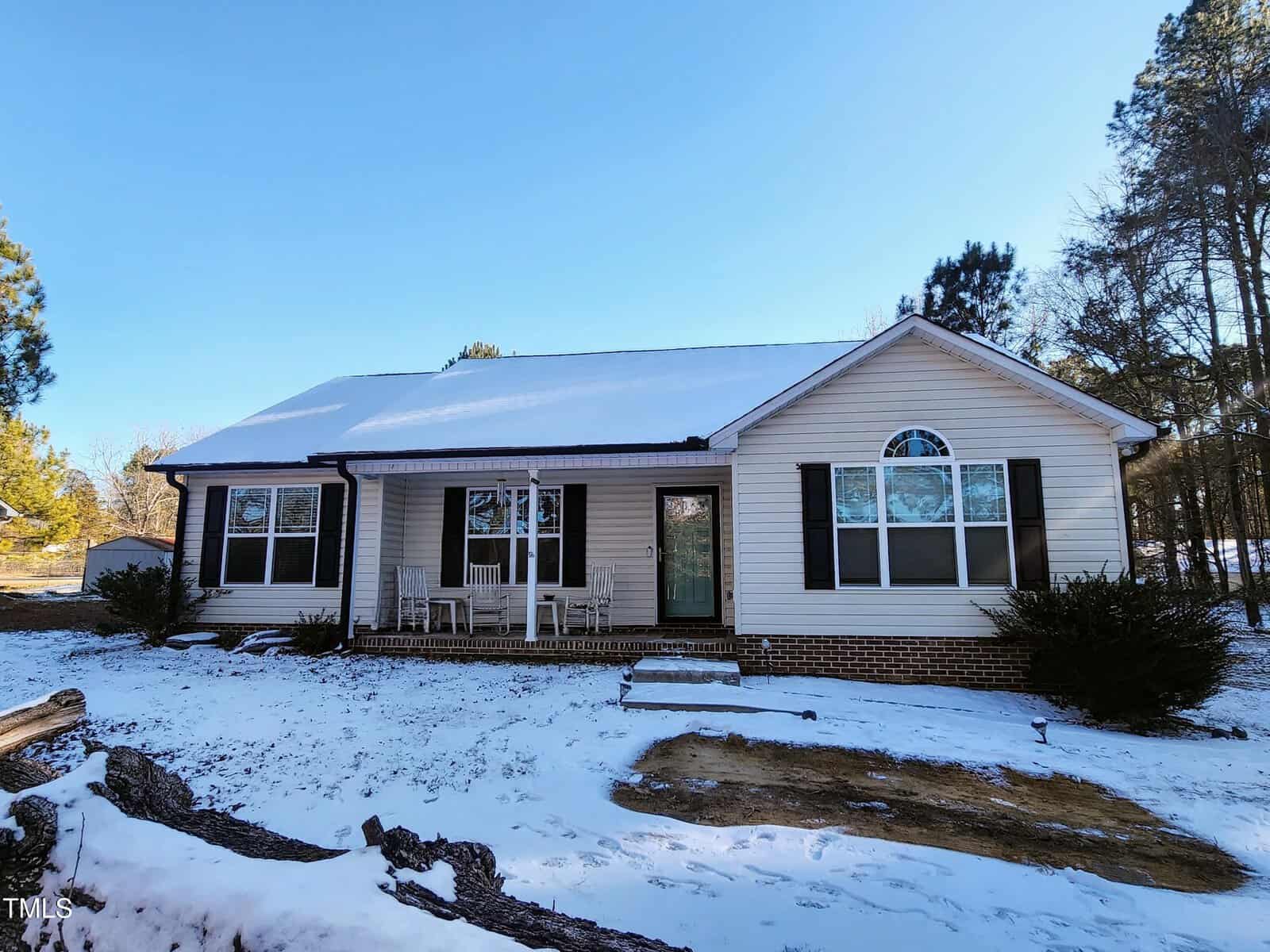 Nestled on 4036 Belltown Road, this charming single-story house with light-colored siding is surrounded by a blanket of snow. It features a welcoming front porch with white chairs and a glass door. Tall trees flank each side under the clear blue sky, embodying the serene beauty of Oxford.