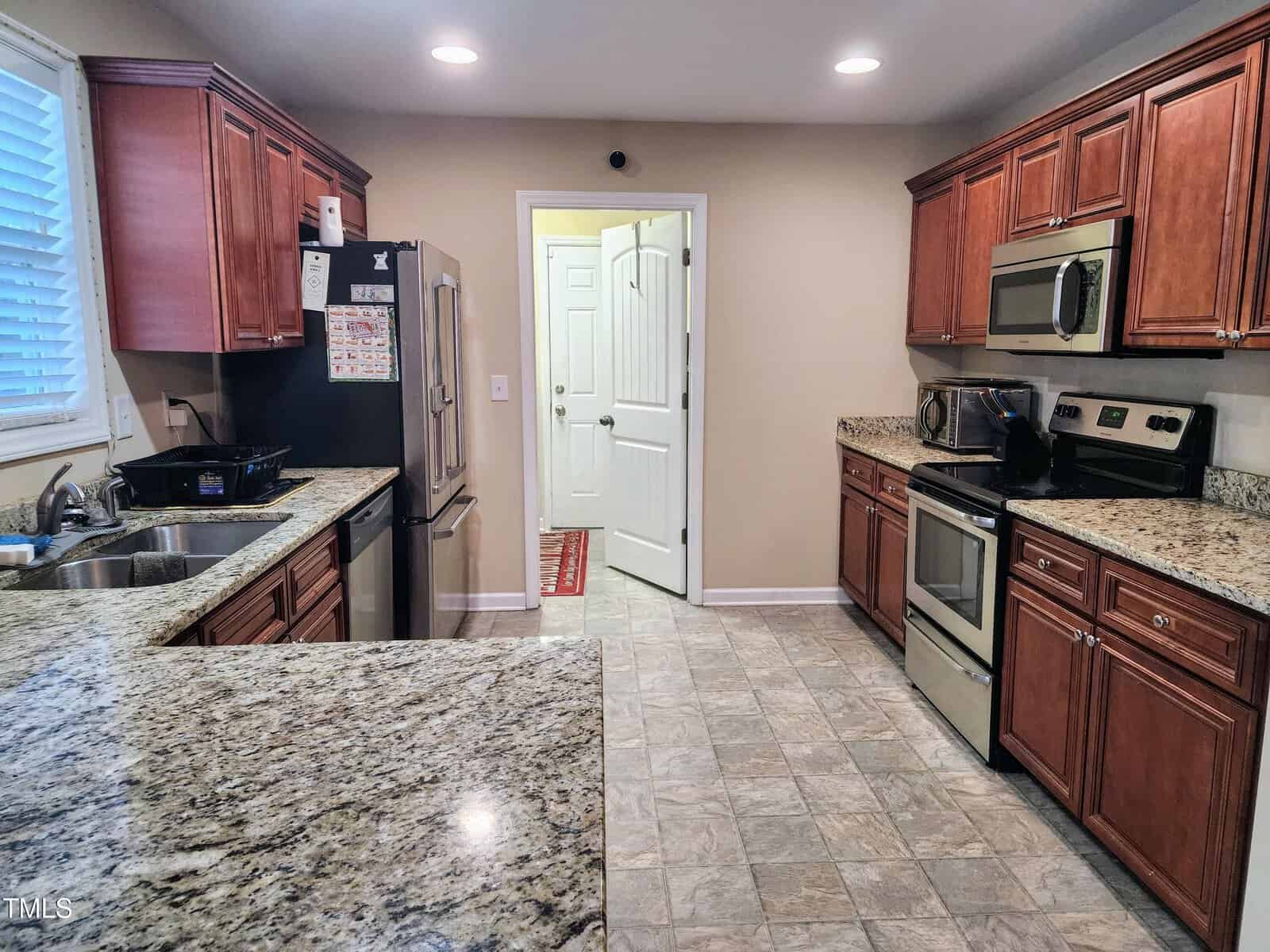 A kitchen with granite countertops, wooden cabinets, and stainless steel appliances including a refrigerator, microwave, and oven. A sink is on the left, and an open door leads to another room. The floor is tiled in a light pattern.