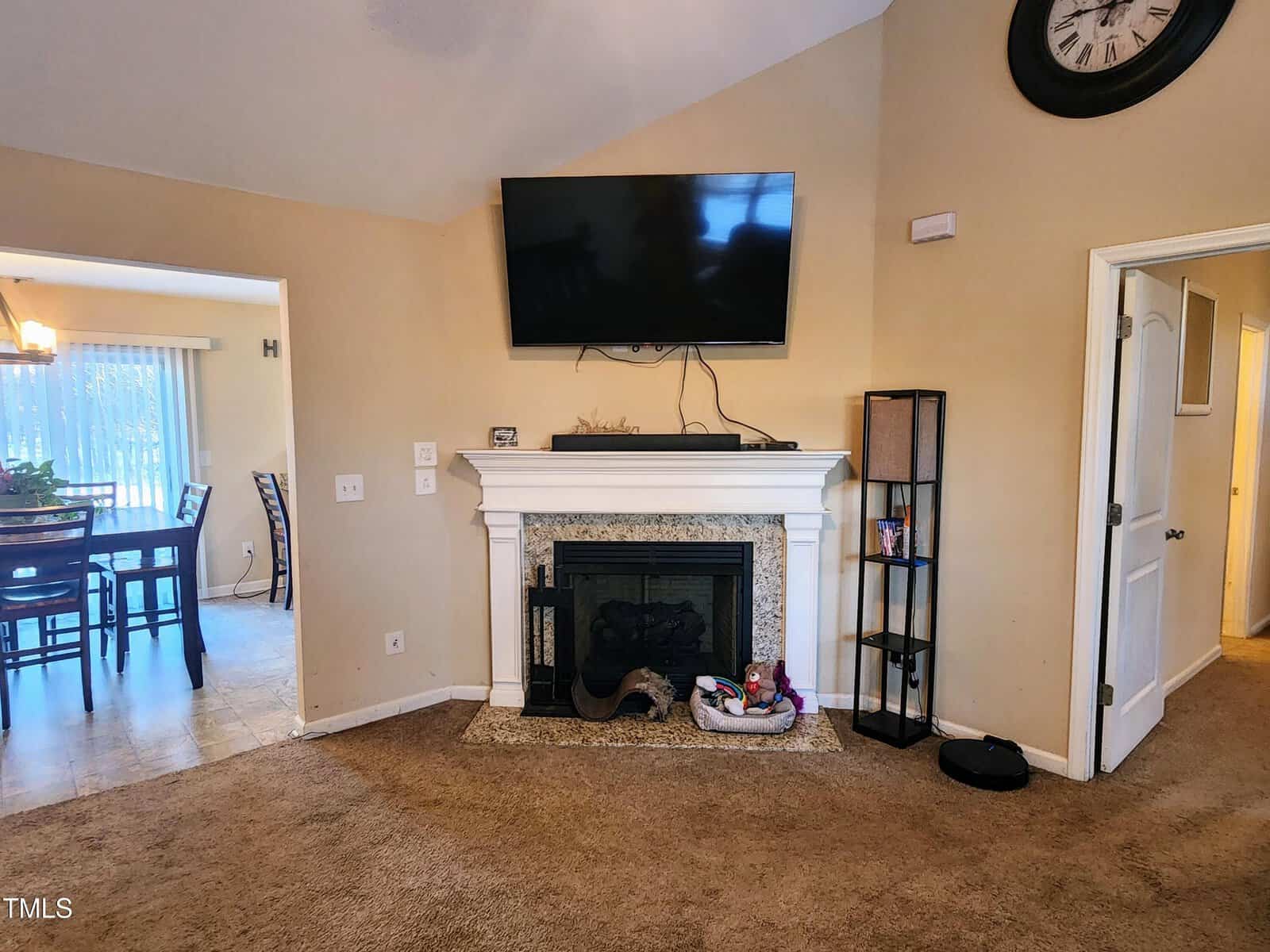 Living room with a wall-mounted TV above a fireplace, surrounded by a white mantel. To the right, a tall, narrow shelf holds a few items. A doorway leads to another room. Dining area with a table and chairs is visible through an opening on the left.