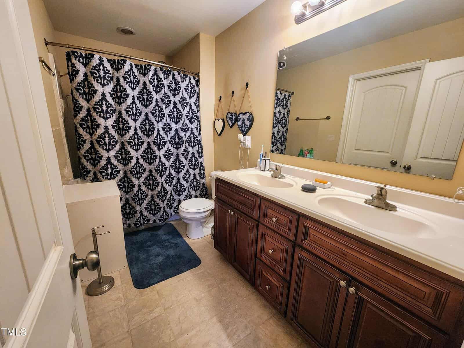 A bathroom with a dual-sink wooden vanity and a large mirror. The shower has a patterned curtain. There's a toilet near the shower and two heart-shaped decorations on the yellow wall. The floor is tiled, and a blue rug is near the shower.