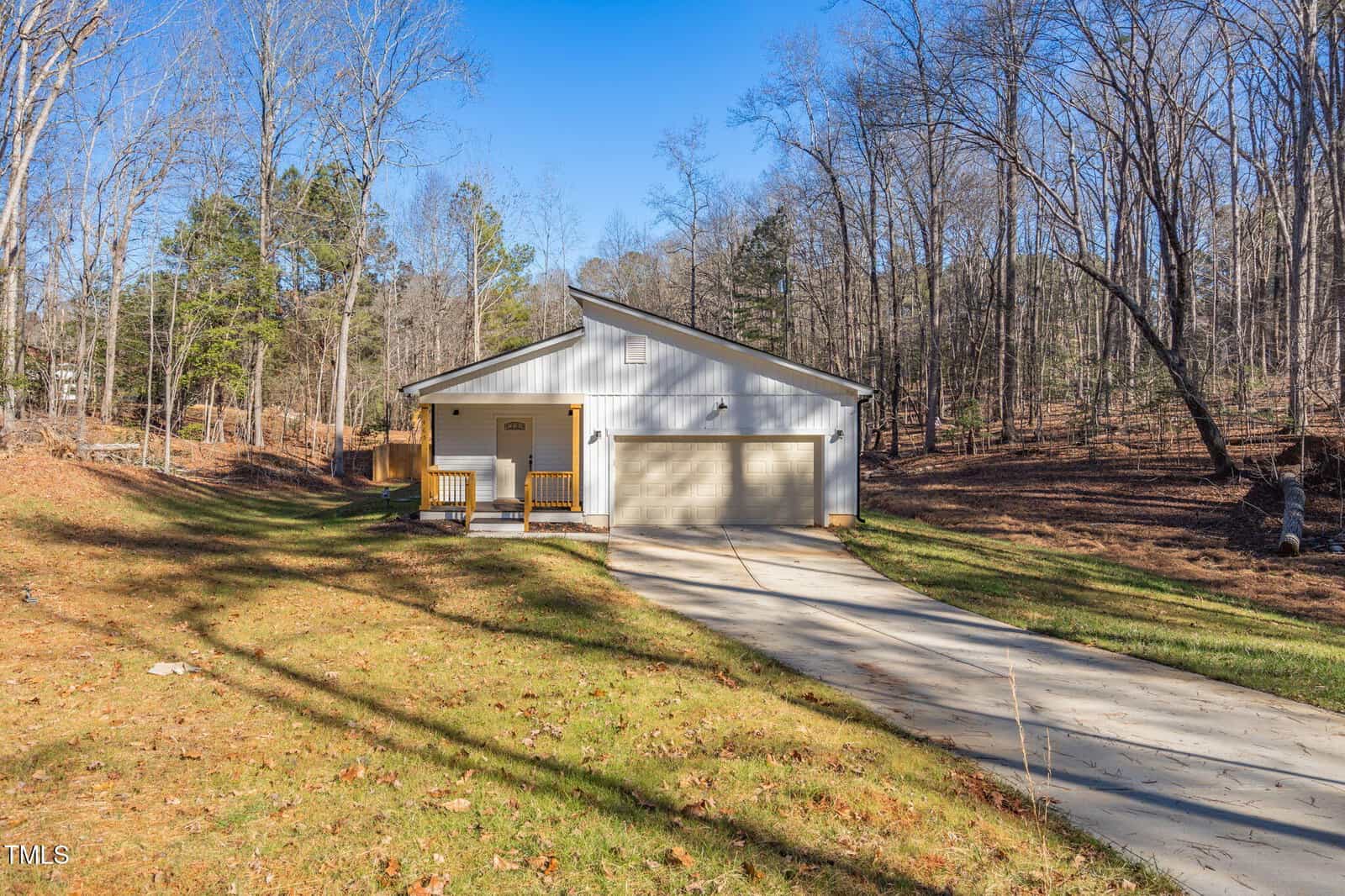 A single-story white house with a two-car garage and a covered front porch sits at the end of a concrete driveway, surrounded by trees and a grassy yard under a clear blue sky.