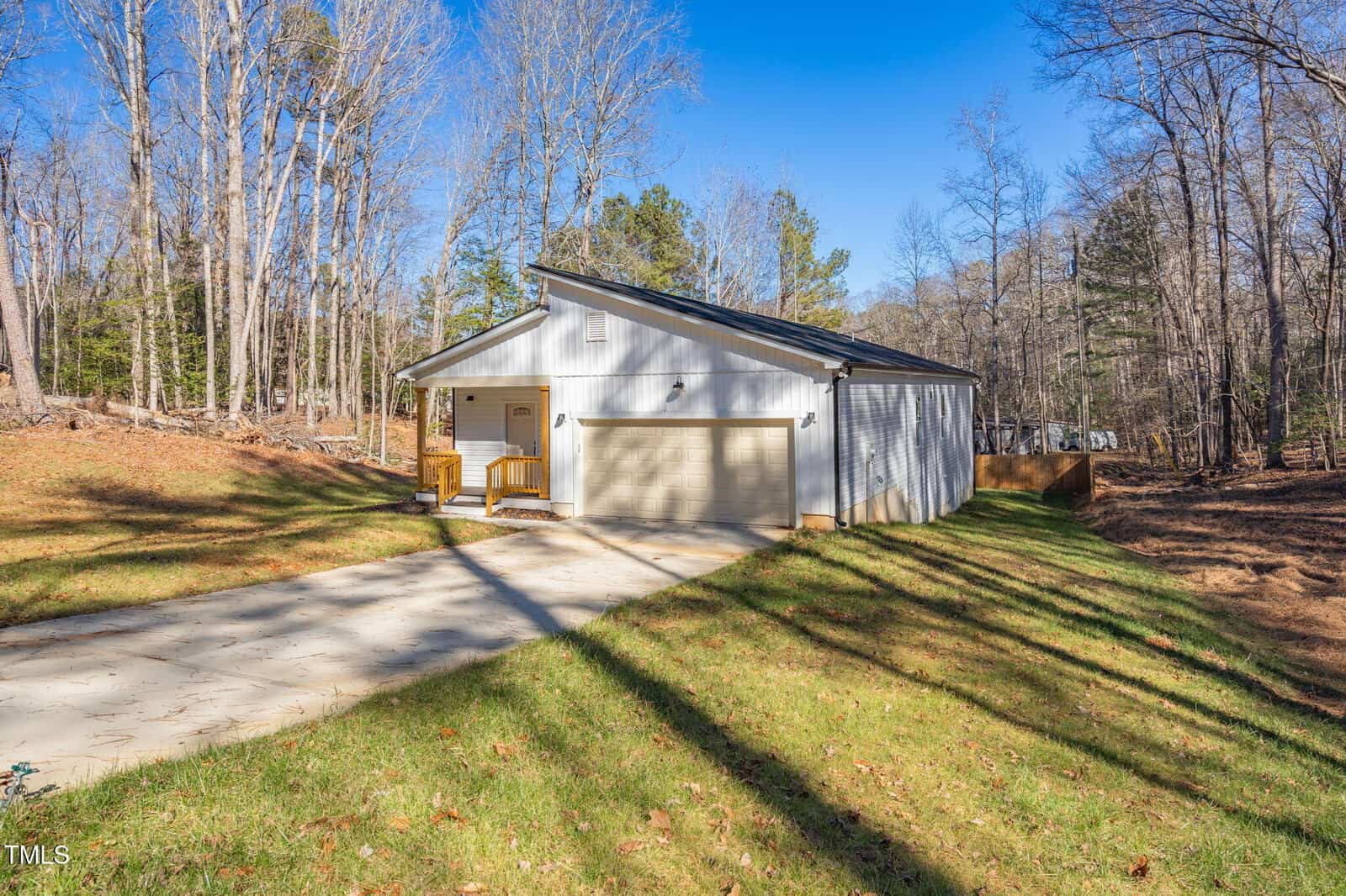 A white single-story house with a two-car garage sits at the end of a concrete driveway, surrounded by grass and trees under a clear blue sky. Two wooden chairs are on the front porch.