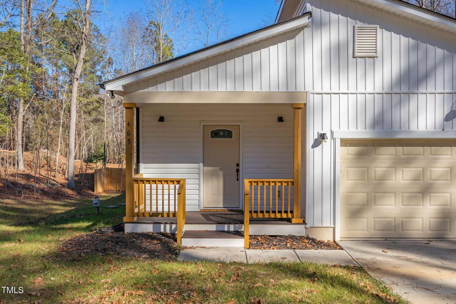 A small front porch with wooden railings and steps leads to a beige front door on a white house with vertical siding. The porch is next to a beige garage door, surrounded by trees and a lawn.