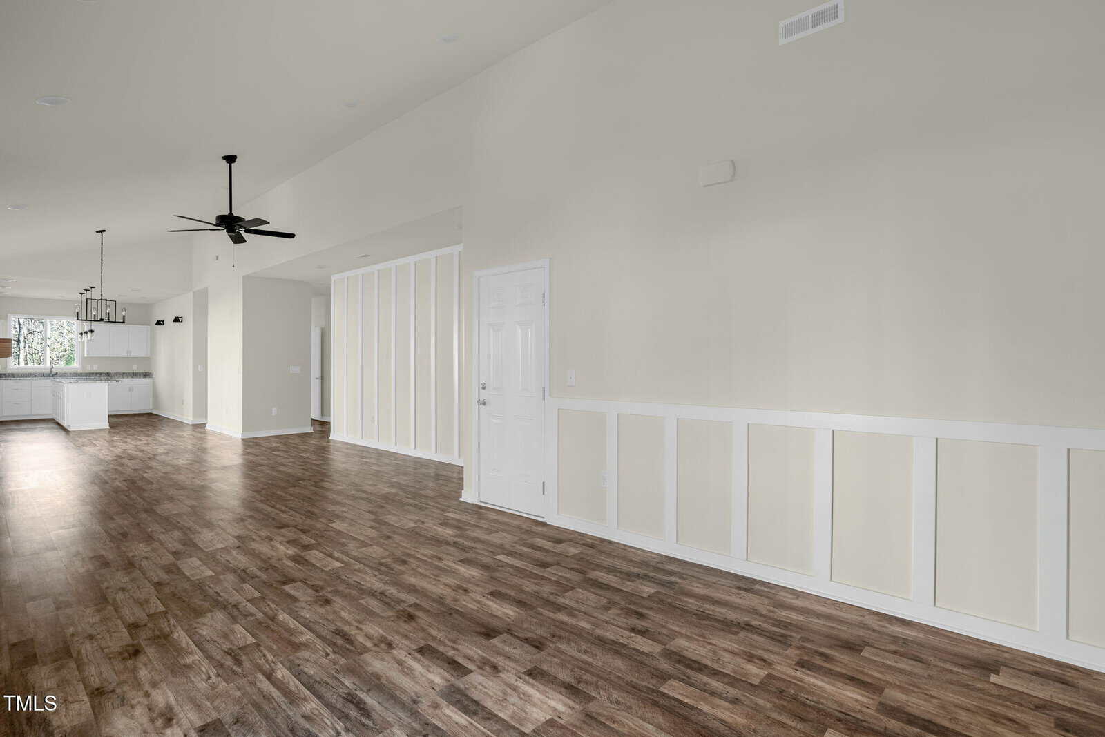 A spacious, empty room with light-colored walls, wood flooring, white trim, a ceiling fan, and a view into a kitchen area with white cabinets and pendant lighting.