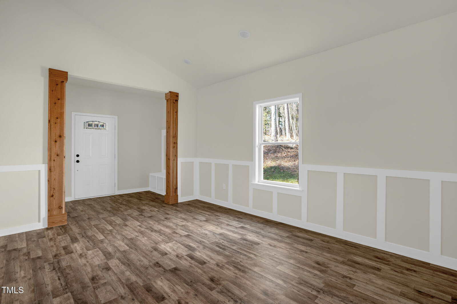 A bright, empty room with light beige walls, white trim, and wood-look flooring. There is a window on the right, a white front door with a small window, and two wooden columns framing a doorway.