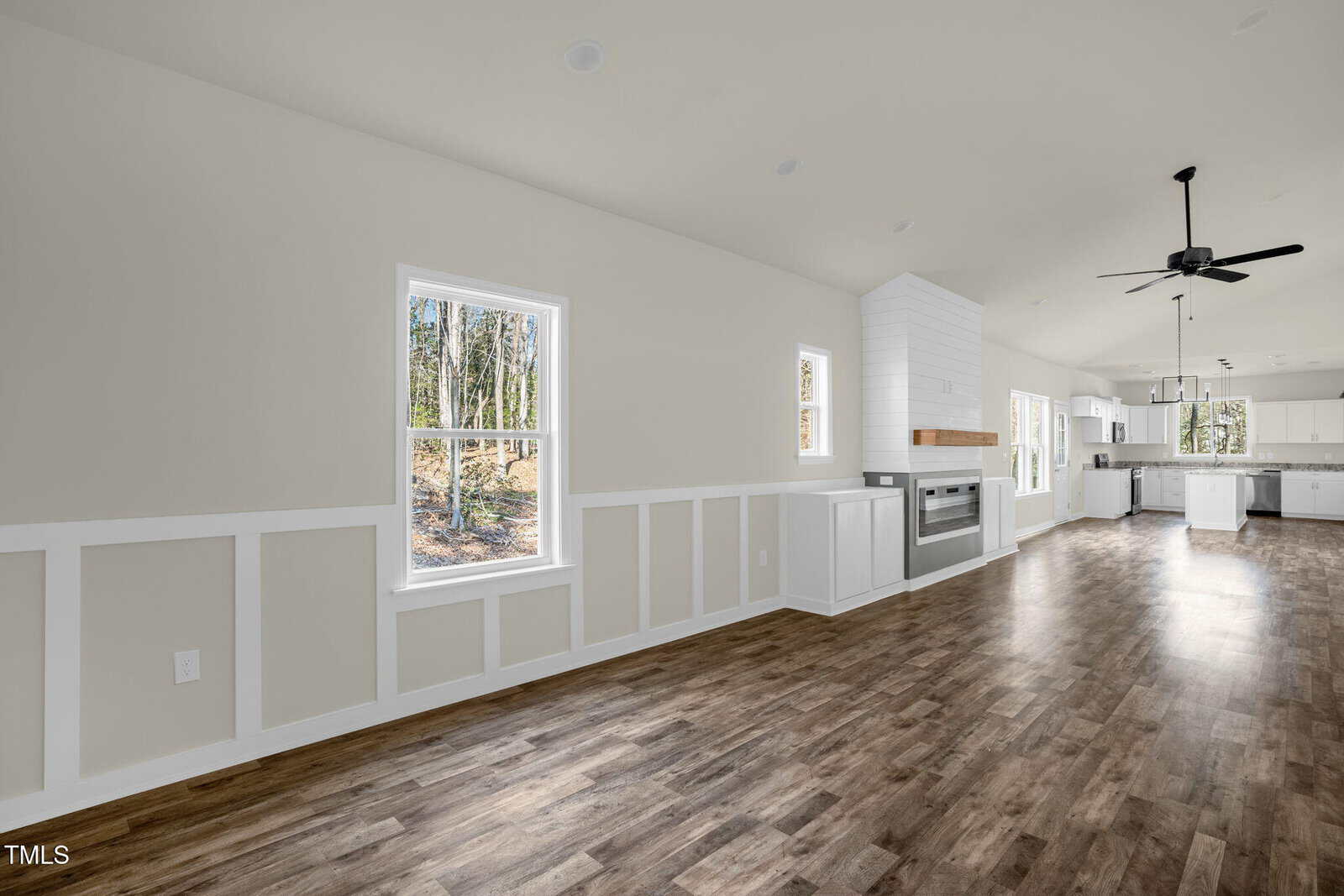 Bright, empty room with wood flooring, white paneled walls, large windows, a fireplace with a white mantel, and an open layout connecting to a modern kitchen and dining area. Ceiling fan and recessed lights are visible.