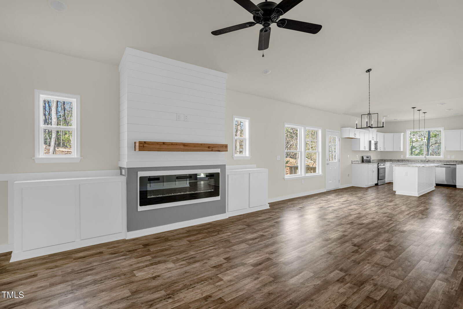 Bright open-concept living space with wood floors, a white shiplap fireplace, ceiling fan, and large windows. The kitchen with white cabinets and a modern light fixture is visible in the background.