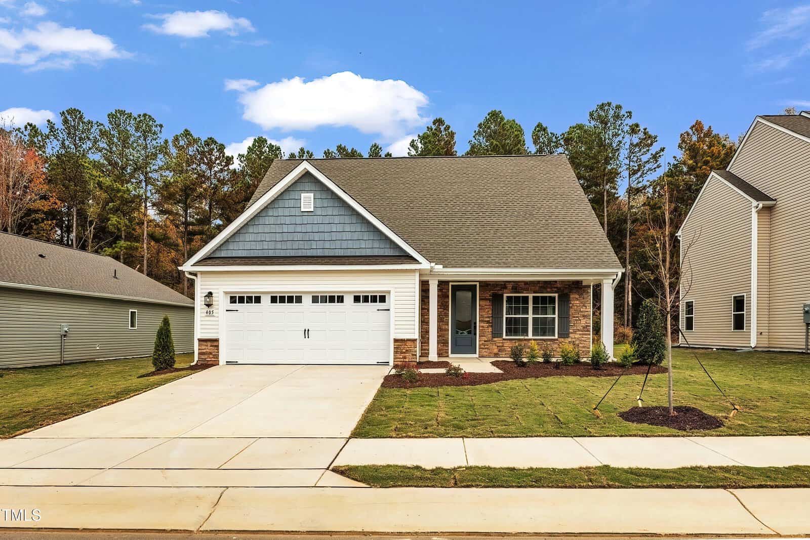 A single-story house with a two-car garage, gray and blue siding, and brick accents. It has a paved driveway, a small front porch, and a neatly landscaped lawn with young trees, set in a suburban neighborhood.
