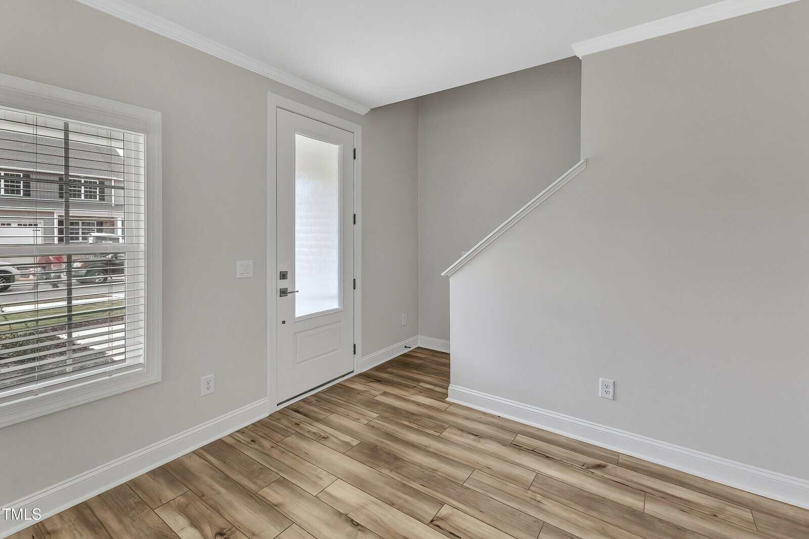 A small, empty room with light gray walls, white trim, wood-look flooring, a window with white blinds, a white door with glass panel, and a staircase with a partial wall.