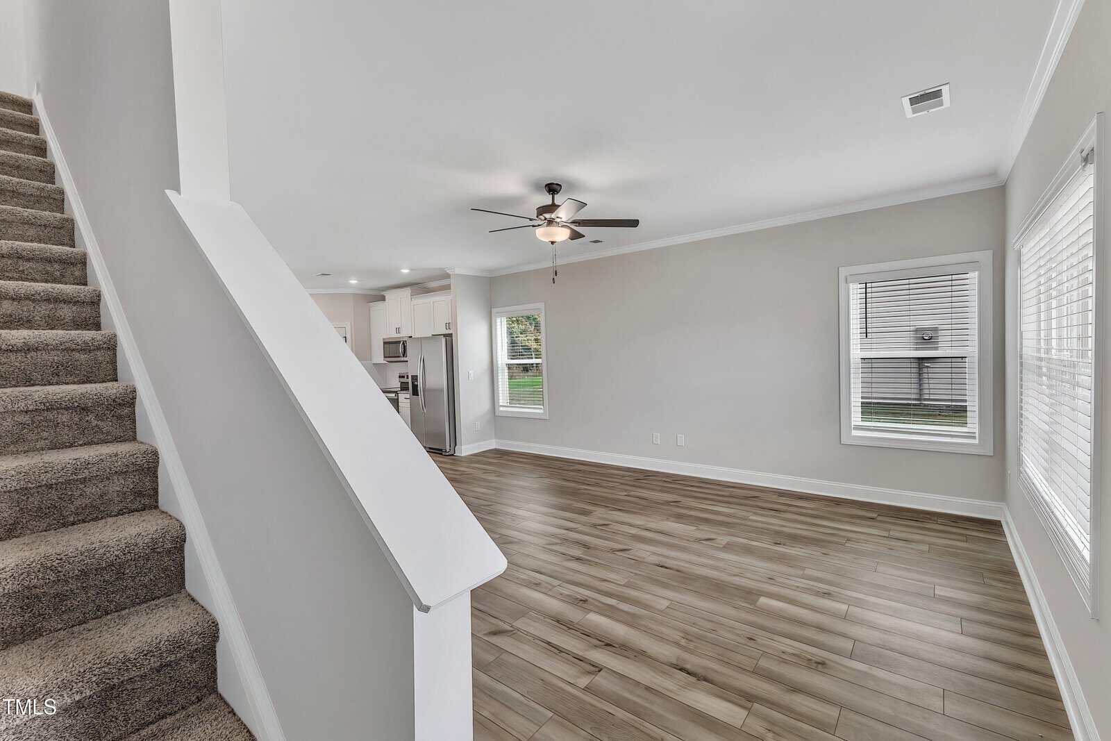 A view of a modern, unfurnished living room with light-colored walls, wood flooring, large windows, a ceiling fan with lights, and a carpeted staircase leading upstairs; the kitchen is visible in the background.