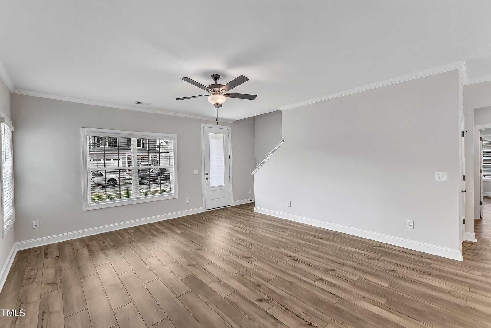 A spacious, empty living room with light gray walls, wood-look flooring, a ceiling fan with lights, a large front window, and a white door next to a short staircase.