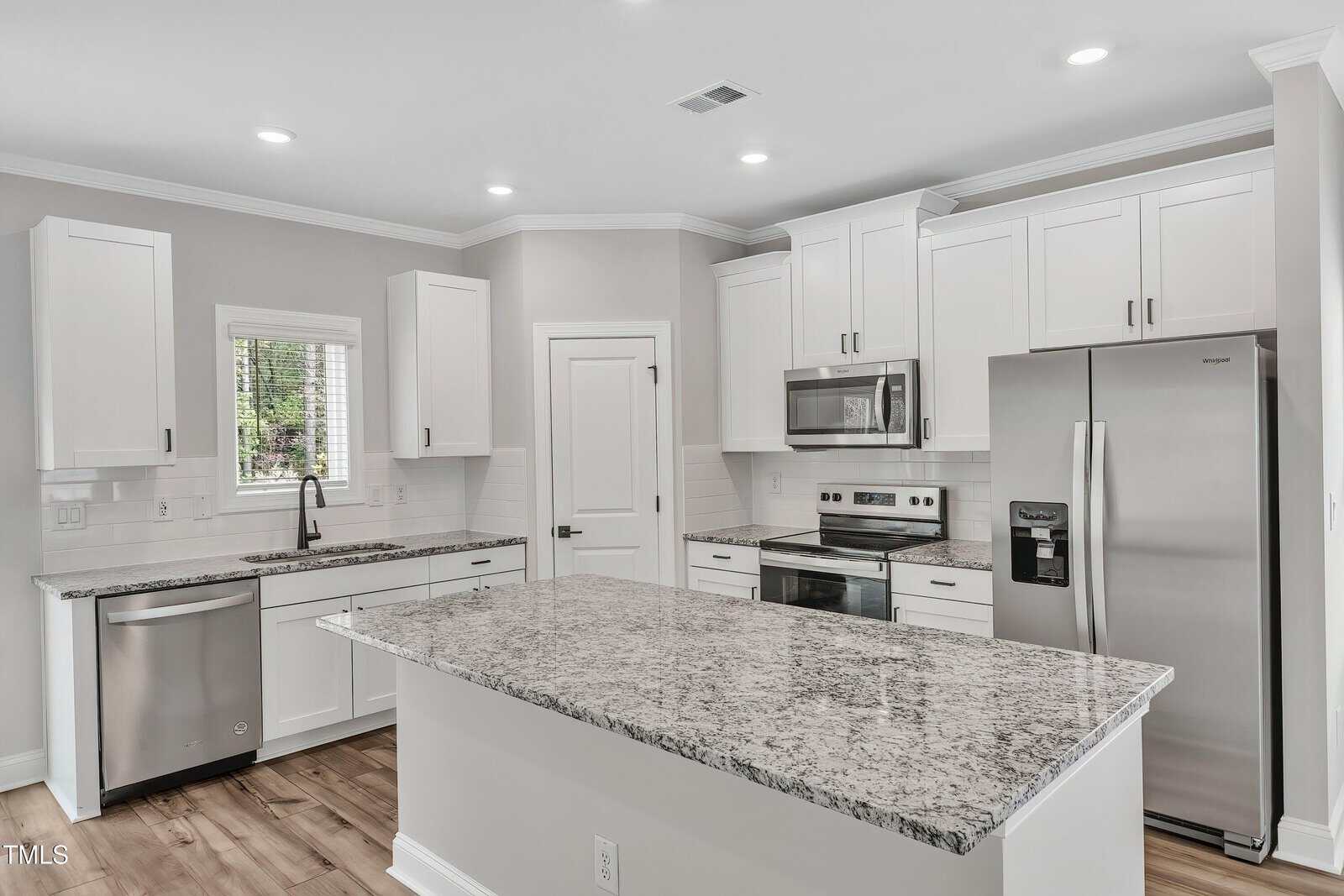 Modern kitchen with white cabinets, stainless steel appliances, granite countertops, an island in the center, light wood flooring, and a window above the sink letting in natural light.