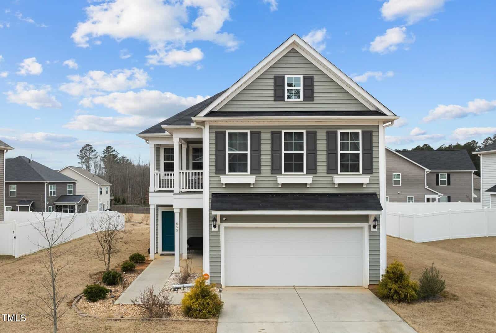 A two-story suburban house with gray siding, black shutters, a covered front porch, and a two-car garage. The yard is mostly brown grass with minimal landscaping, and a white fence surrounds the property.