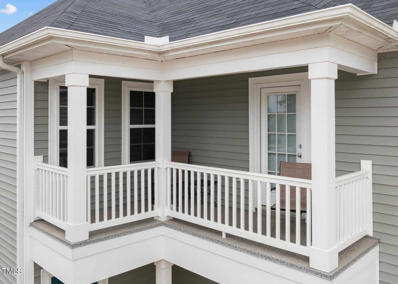 A small covered balcony with white railings and columns, gray siding, two windows, a glass door, and two outdoor chairs.
