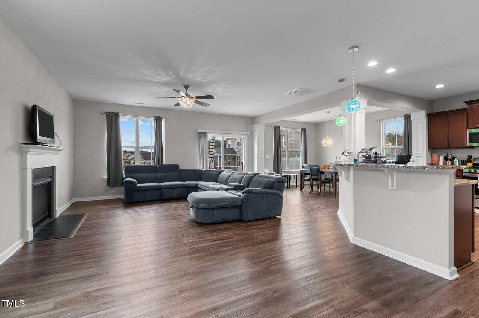 Open-concept living area with wood floors, a sectional sofa, a fireplace with a wall-mounted TV, and a kitchen with pendant lights, granite countertops, and stainless steel appliances. Large windows provide natural light.
