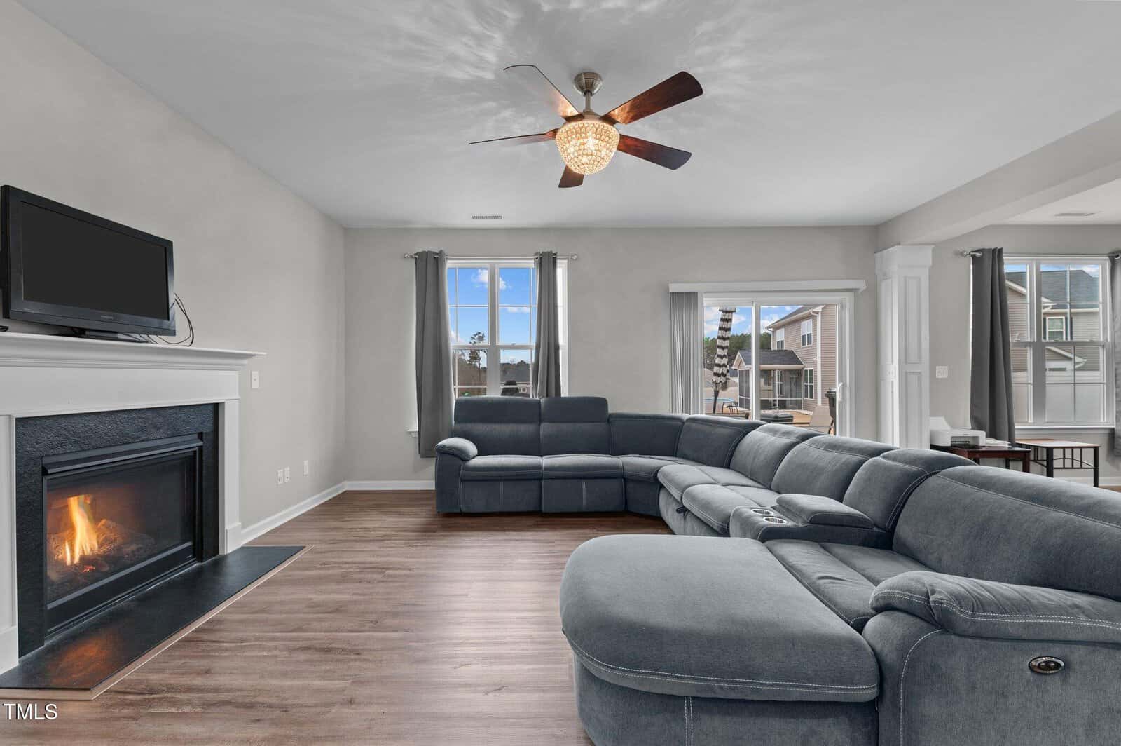 A modern living room with wood floors, a blue sectional sofa, a fireplace with a TV above it, large windows with gray curtains, and a ceiling fan with a light fixture.