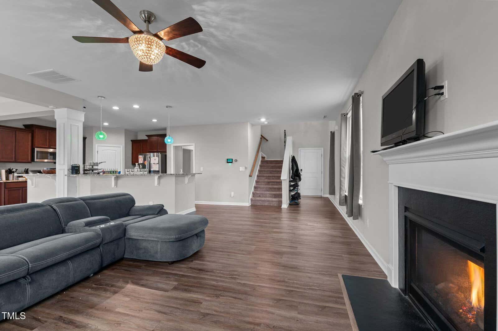 Modern living room with a gray sectional sofa, fireplace, wall-mounted TV, and open kitchen. Wooden floors, ceiling fan, and staircase leading upstairs. Green and blue pendant lights hang over the kitchen counter.