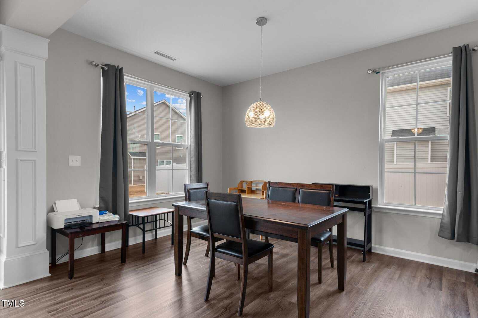 Dining area with dark wood table and four chairs, two windows with dark curtains, a pendant light, a printer on a side table, a bench, and a piano against the wall, all on wood flooring.
