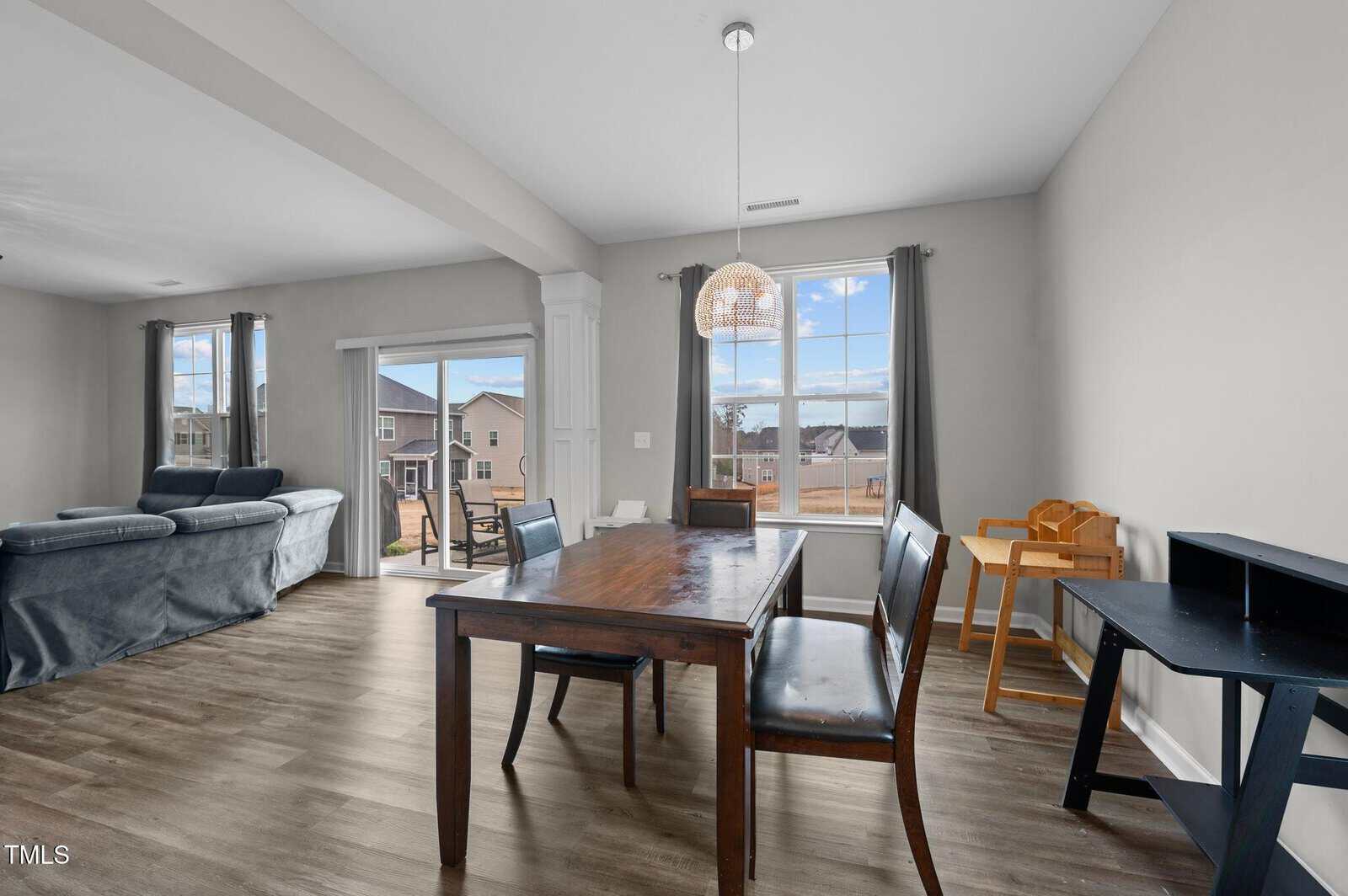 Dining area with a wooden table and four chairs next to large windows. In the background, there’s a living room with couches, hardwood floors, and views of neighboring houses outside.