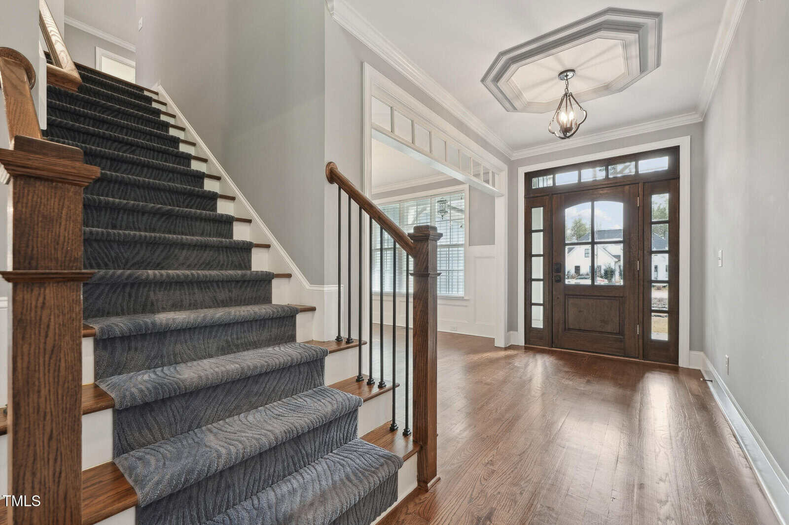 A residential foyer with polished wooden floors and a dark wooden staircase with a blue carpet runner. A modern light fixture hangs from the ceiling, and a wooden front door with large glass panels lets natural light in. White trim accents the walls.