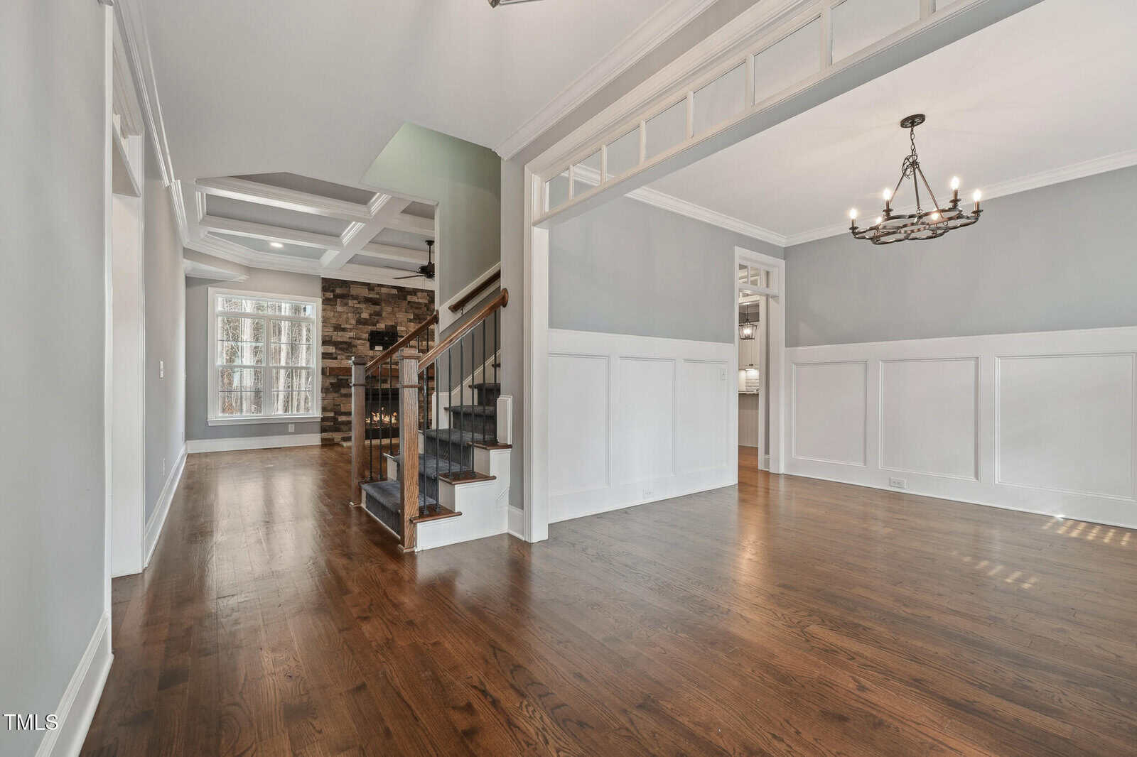 Spacious interior of a modern home featuring a hallway with hardwood floors and a view of a staircase. The walls are light gray with white wainscoting, and a room with a chandelier is visible to the right. A large window and fireplace are in the distance.
