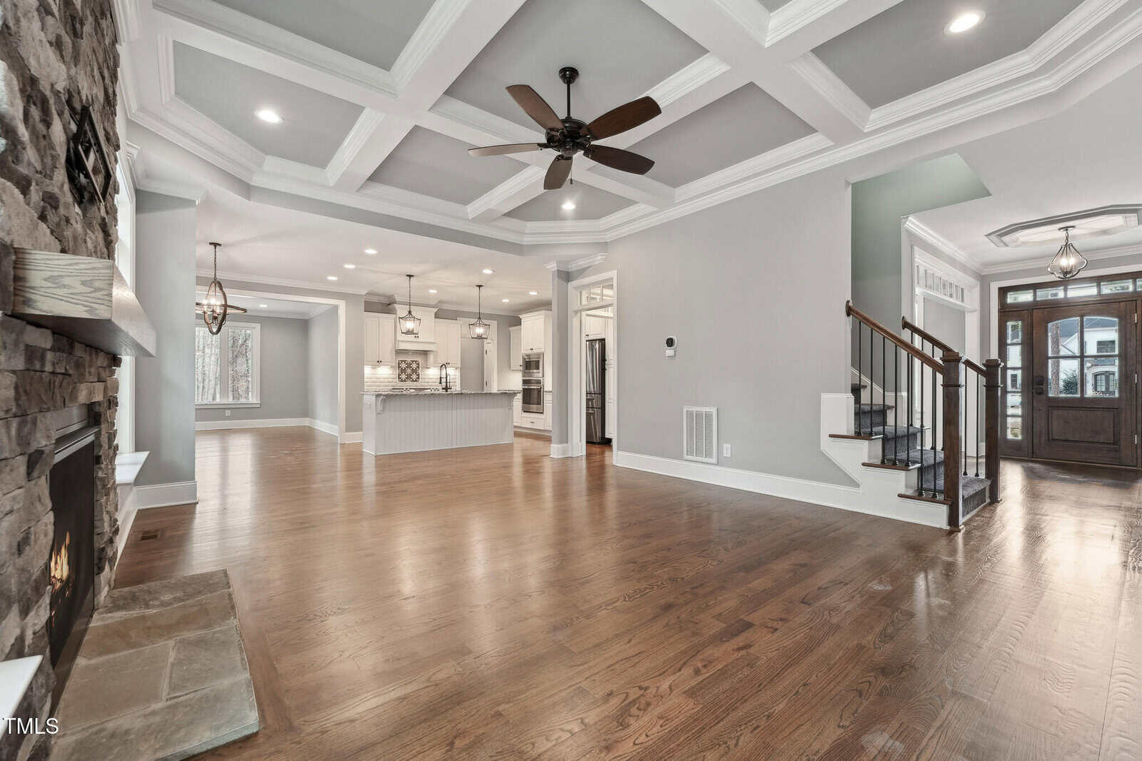 Spacious living area with a wooden floor, stone fireplace on the left, and a coffered ceiling with a ceiling fan. The open floor plan leads to a bright kitchen with white cabinets and an island. A staircase is visible on the right.