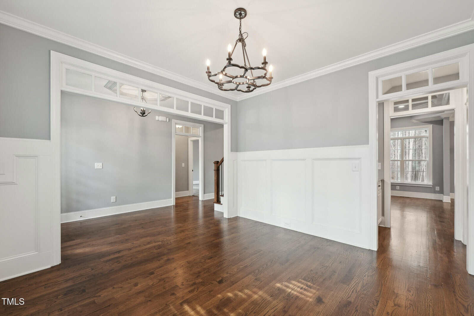 A room with wooden floors and light gray walls featuring white wainscoting. There is a black chandelier on the ceiling and large windows visible through open doorways, providing natural light.