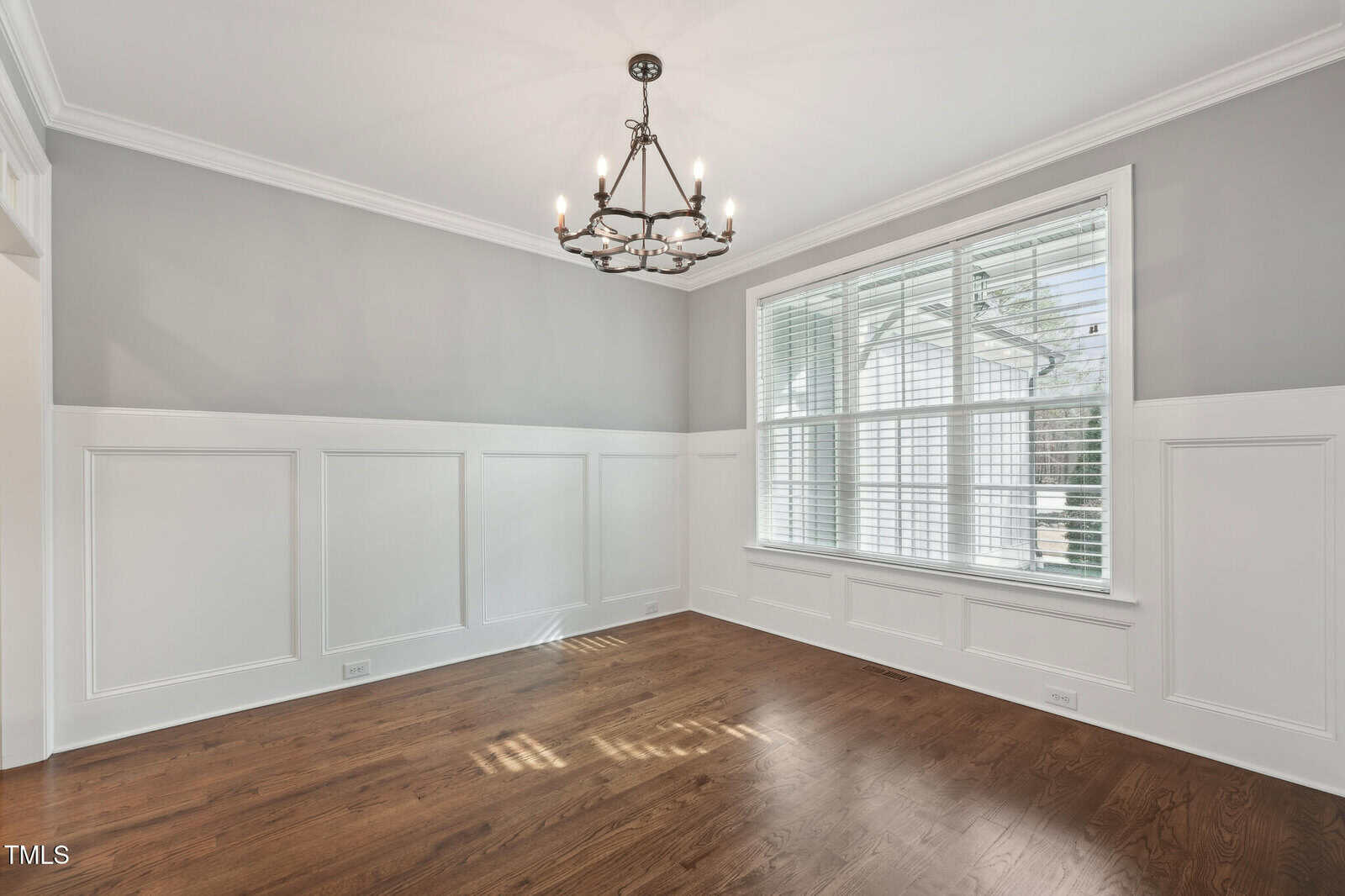 An empty dining room with light gray walls and white wainscoting. A dark metal chandelier hangs from the ceiling. The room has a large window with white blinds, and the floor is wooden.