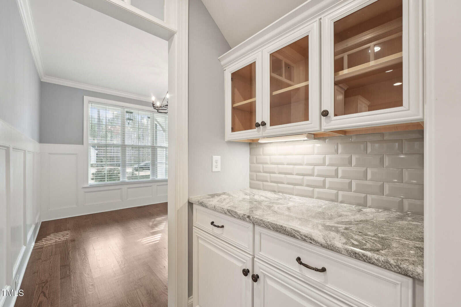 A kitchen area with white cabinets, some with glass doors, and a light gray stone countertop. The backsplash features a light gray subway tile design. Adjacent is a room with large windows and wooden flooring.